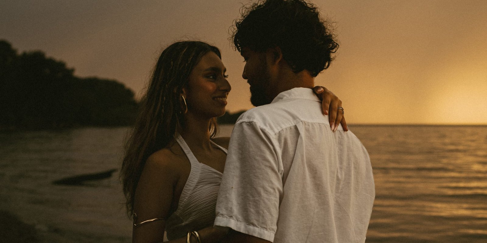 A couple embracing on a lakeshore in warm sunset light, the sky glowing orange behind them
