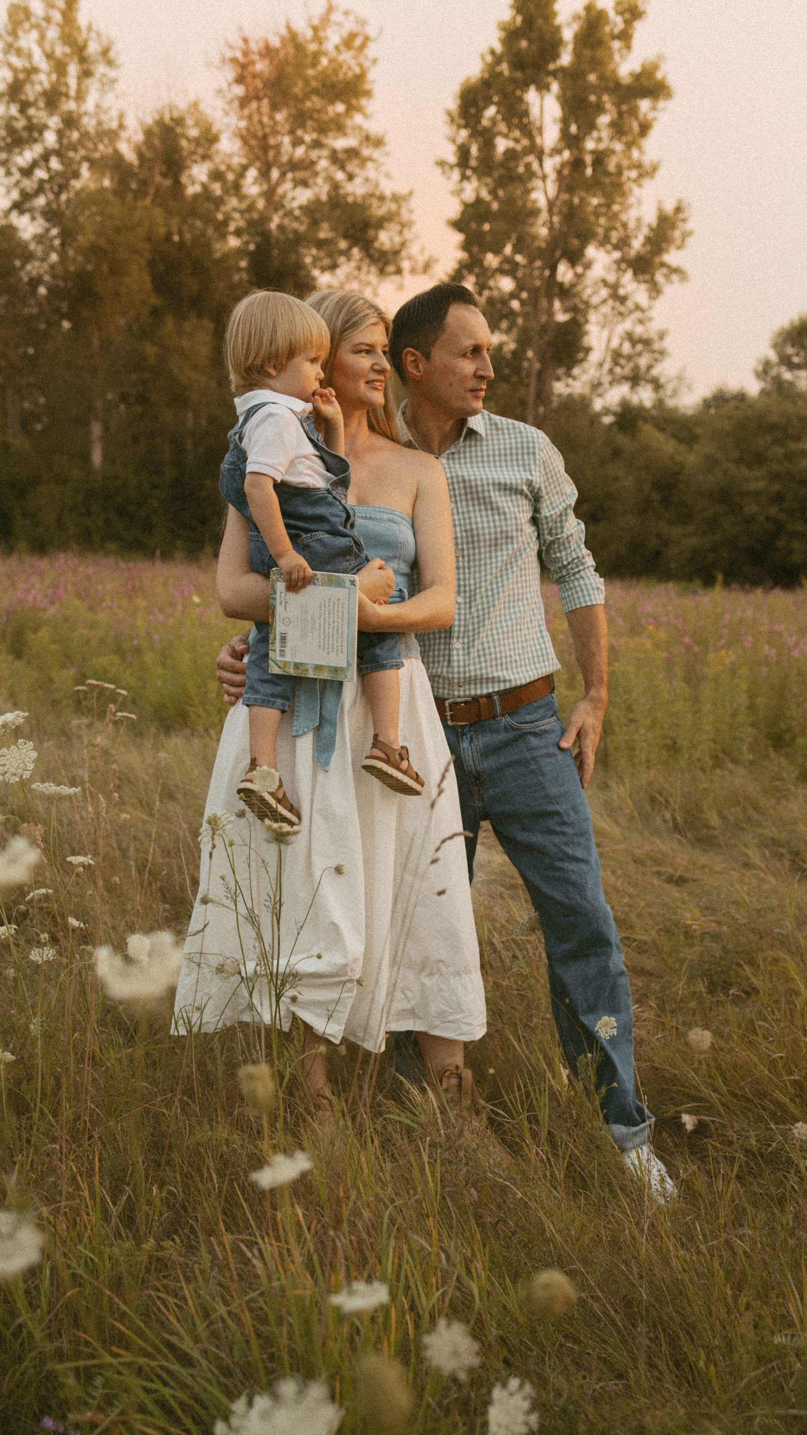 A father holding his young son on his arm in tall grass under a soft pink evening sky, the mother beside them