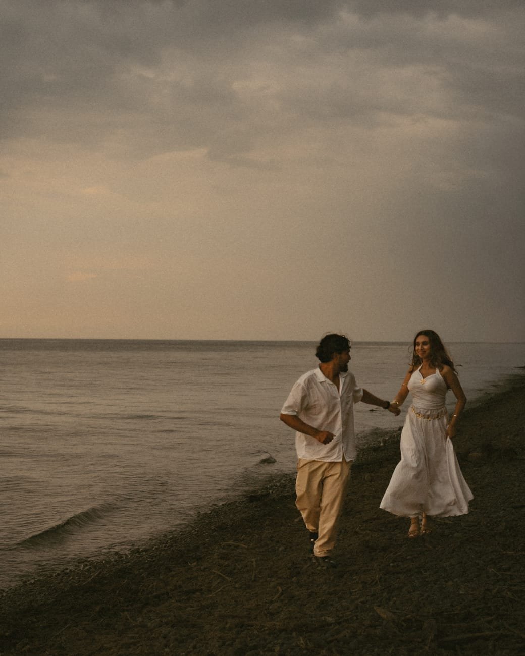 A couple walking hand in hand along the empty shoreline under a wide grey sky