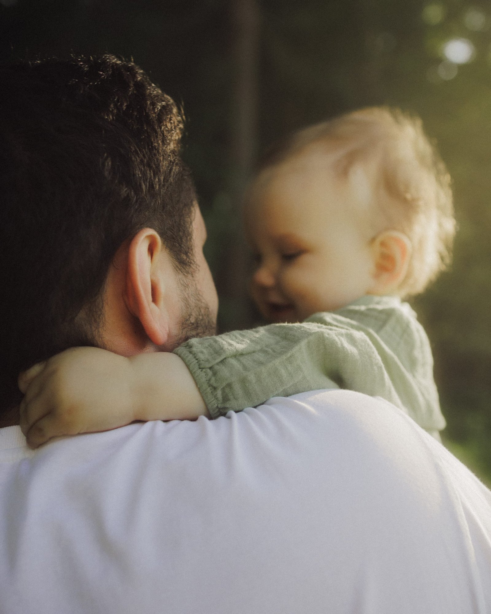 A father lifting his laughing baby overhead beneath tall trees