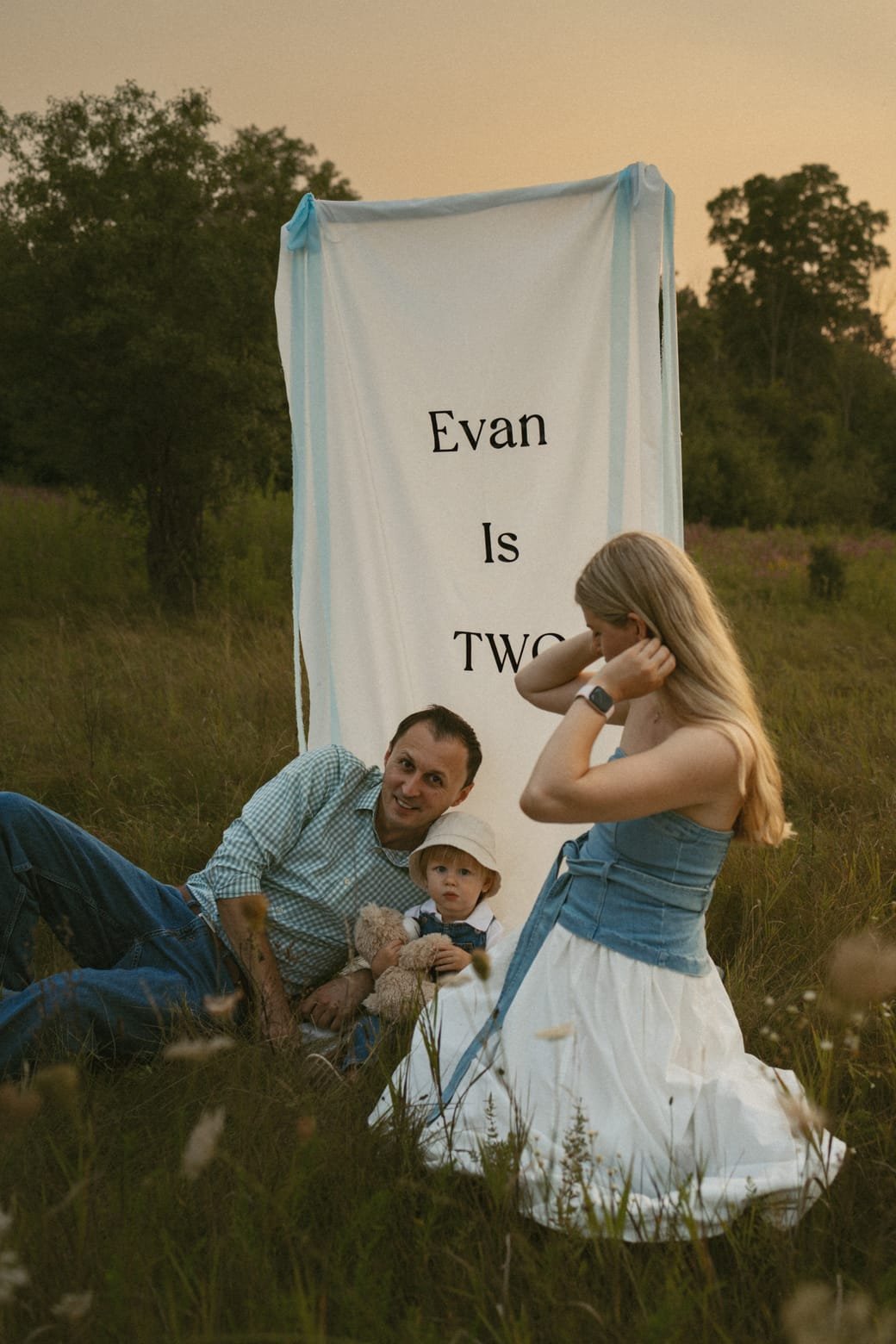 A mother, father and toddler sitting in a wildflower meadow beside a hanging fabric banner at sunset