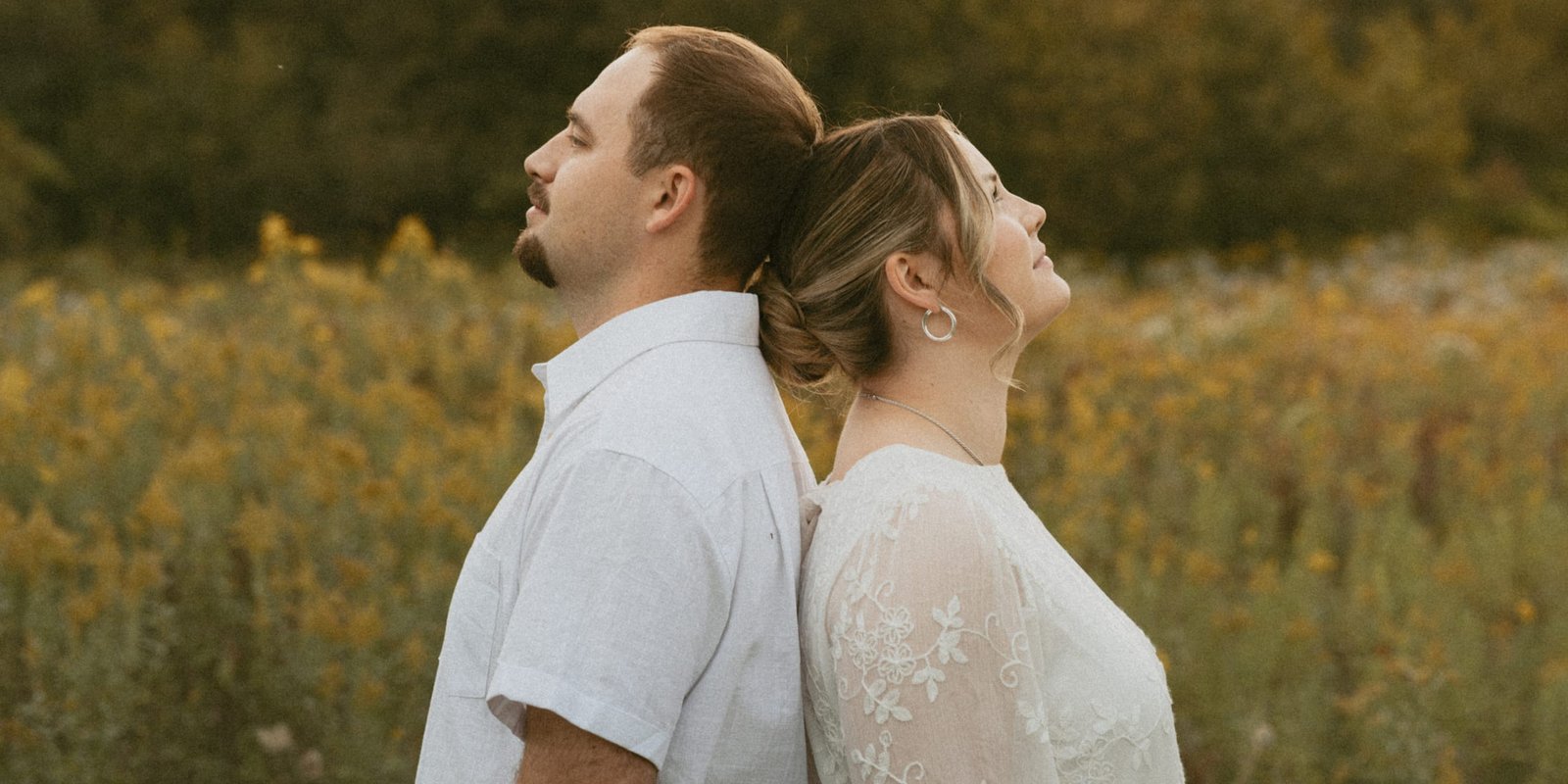 A newly engaged couple standing back to back in a golden field of goldenrod, looking up at the evening sky