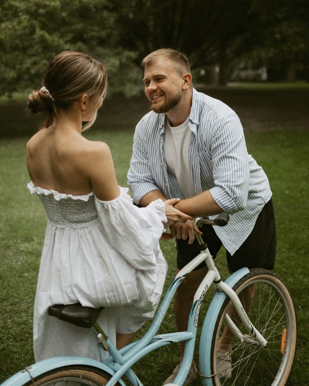 A woman in a white off-shoulder dress steadying a vintage blue bicycle while her partner holds her