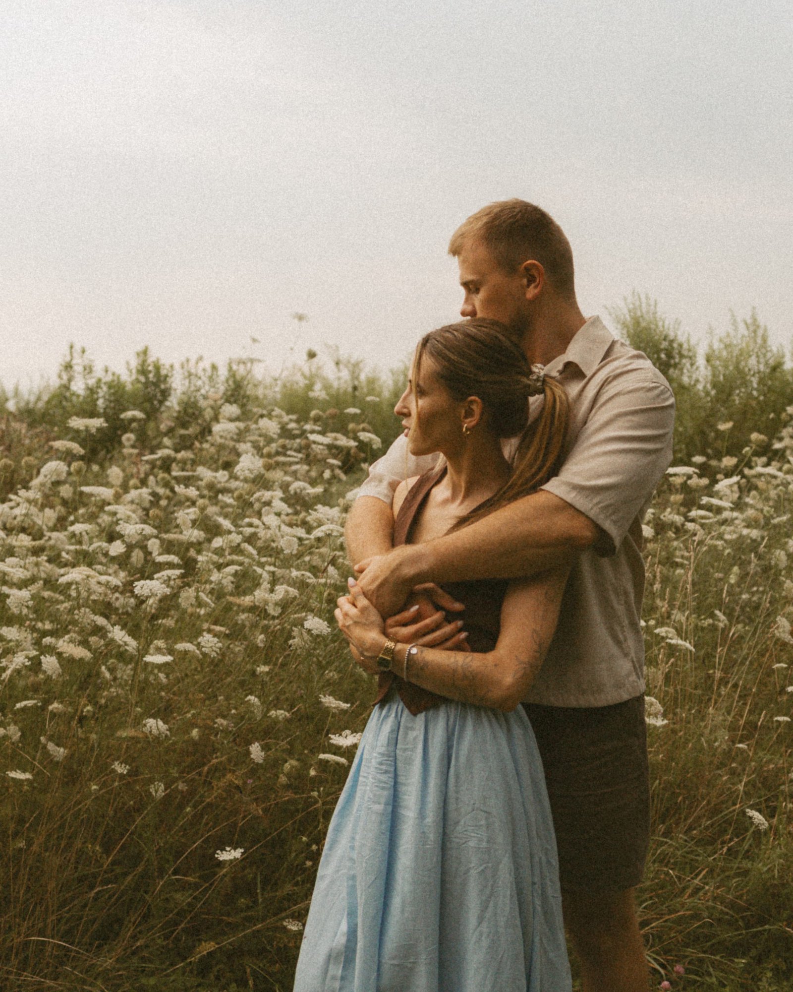 A man holding his partner from behind in a field of Queen Anne's lace, both of them with eyes closed