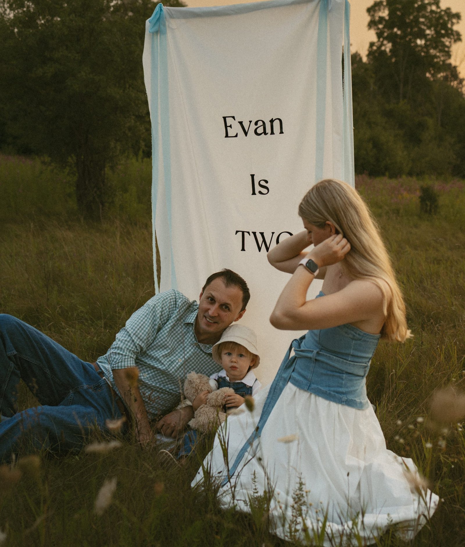 A young family sitting together in a wildflower meadow at sunset beside a hanging linen banner