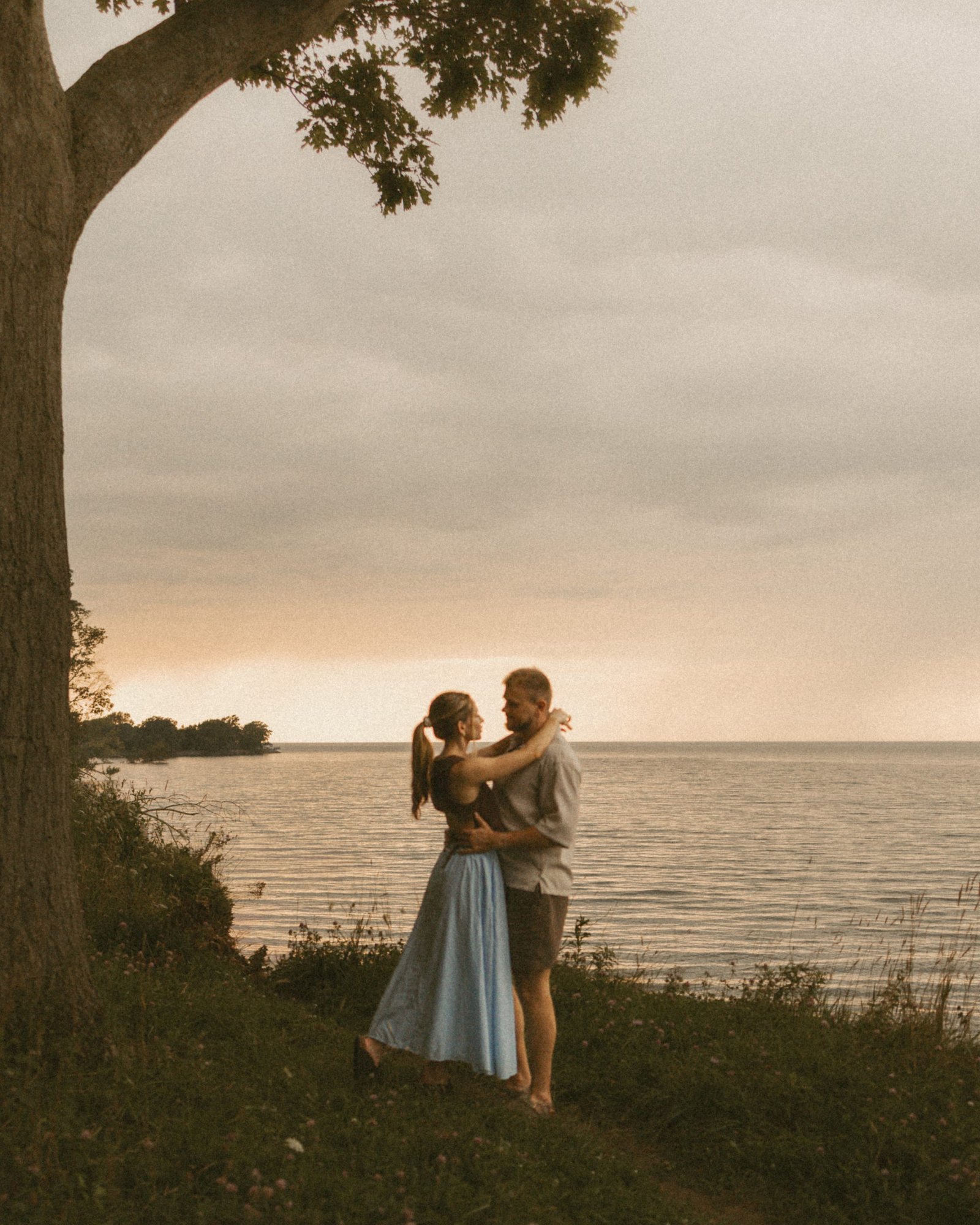 A couple embracing on a grassy bluff beneath a tree, Lake Ontario and a peach-coloured sunset sky behind them