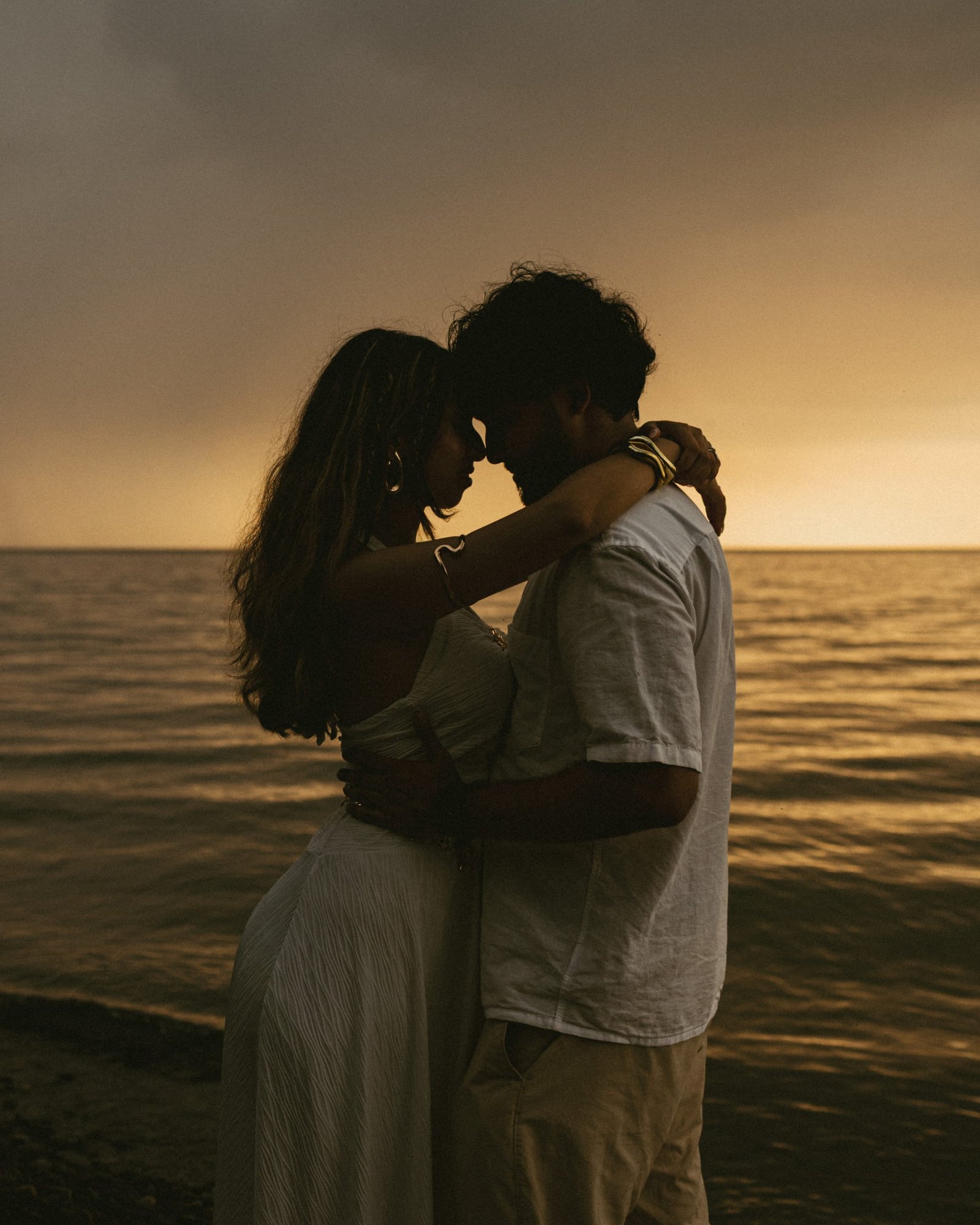 A couple standing close together on a stony lakeshore as the sun sets behind heavy cloud