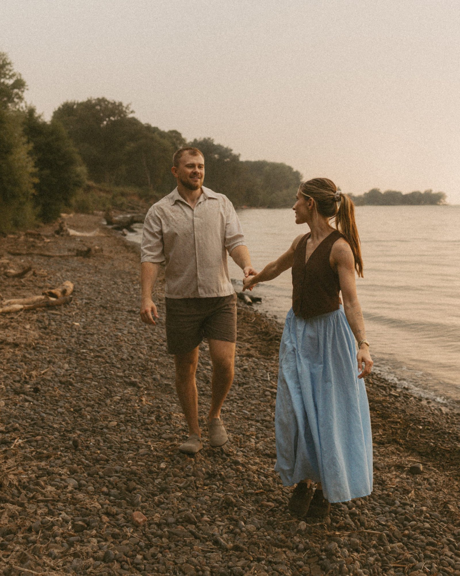 A man leading his partner by the hand along a stony beach, driftwood and the lakeshore trees behind them