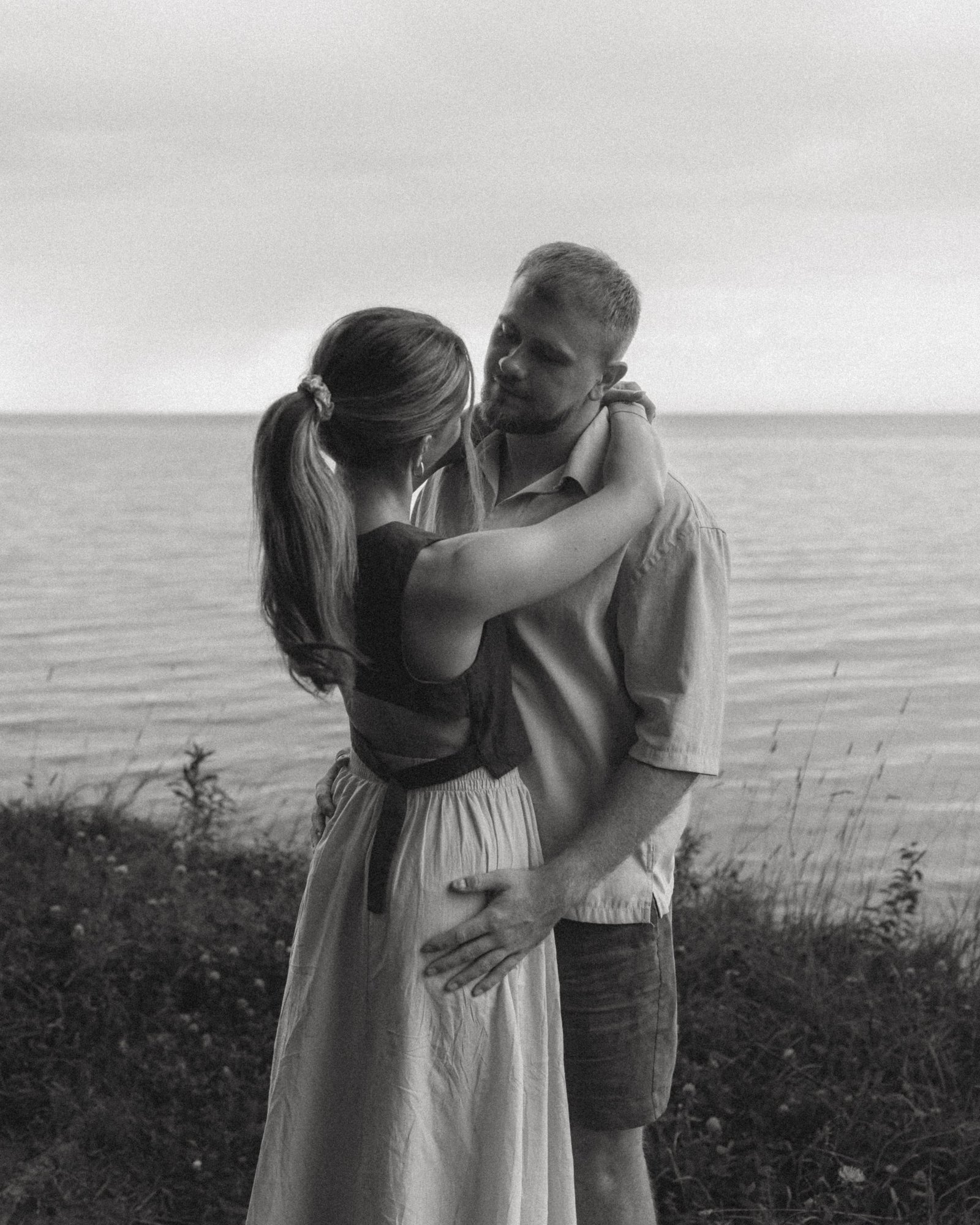 A black and white frame of a couple in a close embrace on the bluff, the flat lake and an overcast sky behind them