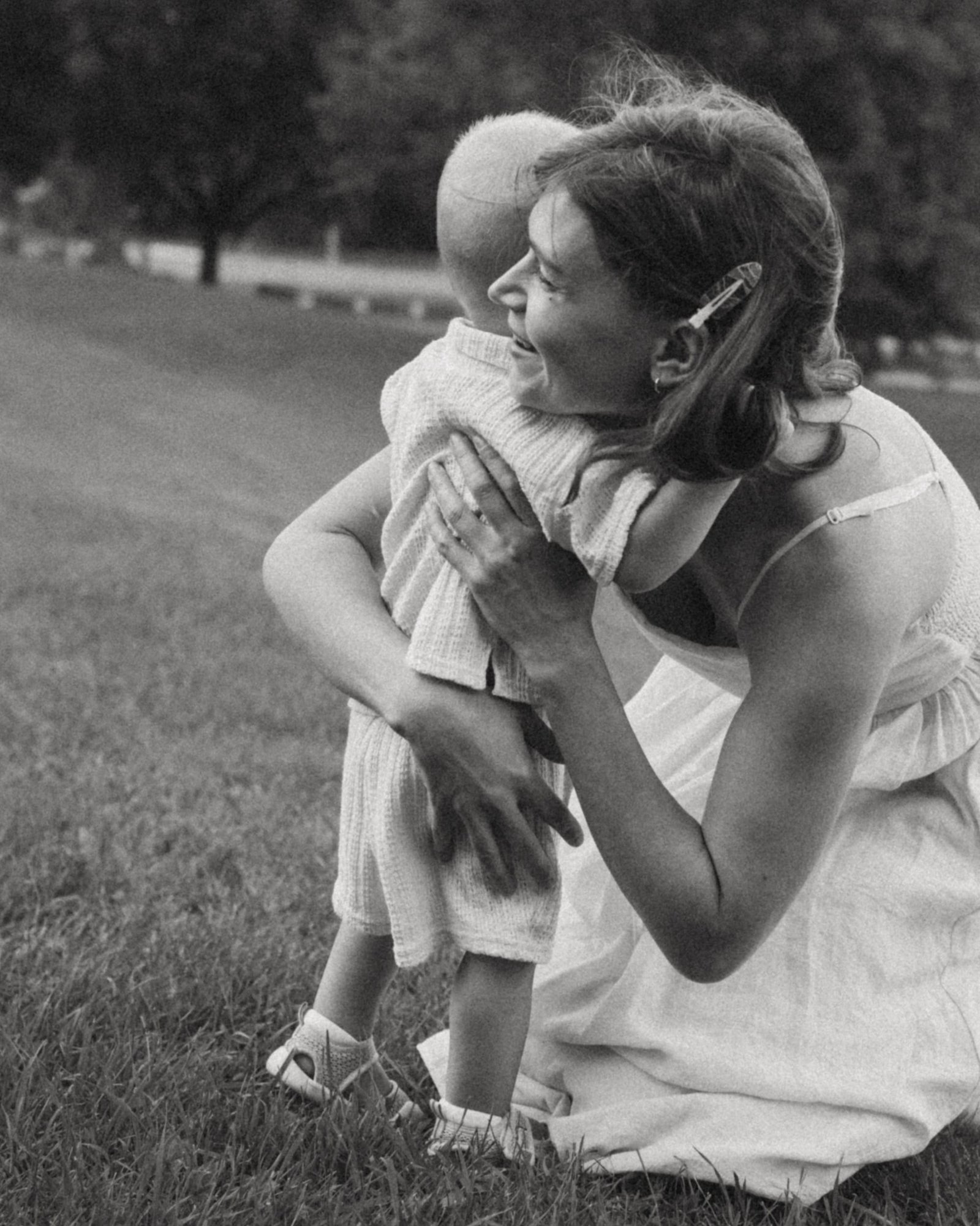 A black and white frame of a mother kneeling in the grass to hug her small son, both of them smiling