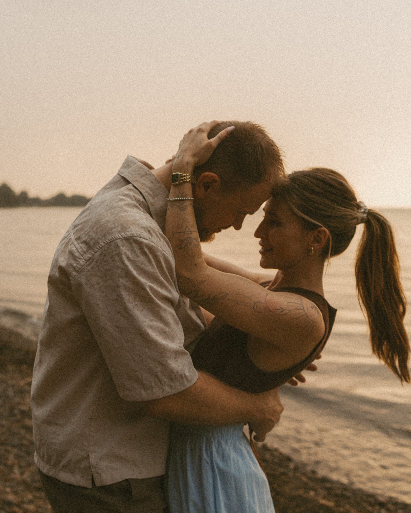 A couple forehead to forehead on the beach at sunset, his hand in her hair, the lake glowing behind them