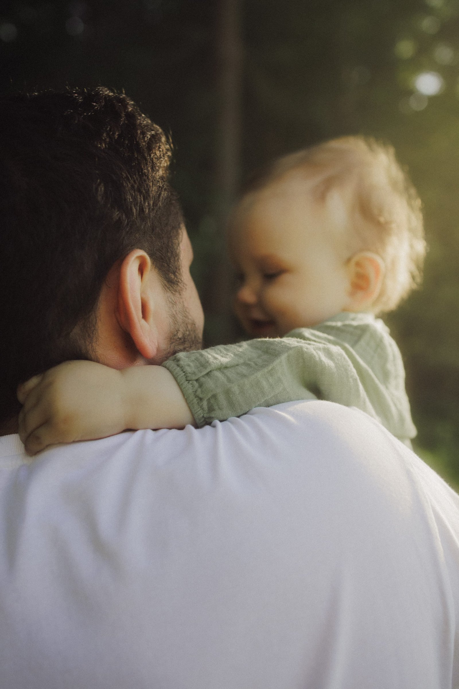 A father lifting his laughing baby overhead beneath tall trees