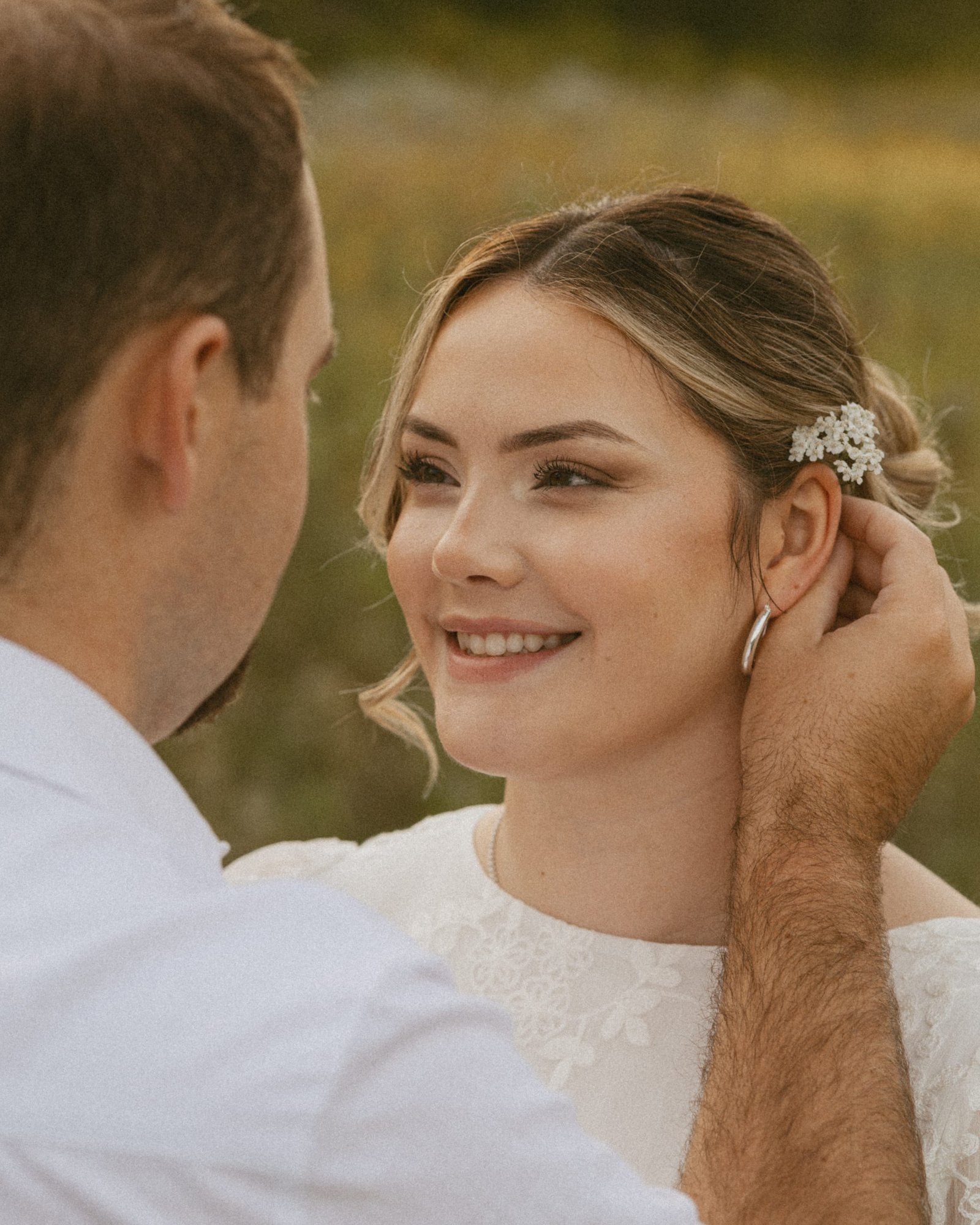 A man tucking a white wildflower behind his fiancée's ear as she smiles up at him