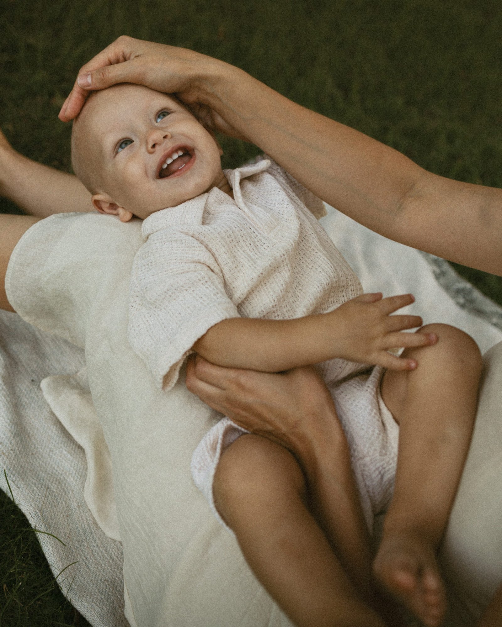 A baby boy lying on his back on a blanket, laughing up at his mother as she strokes his head