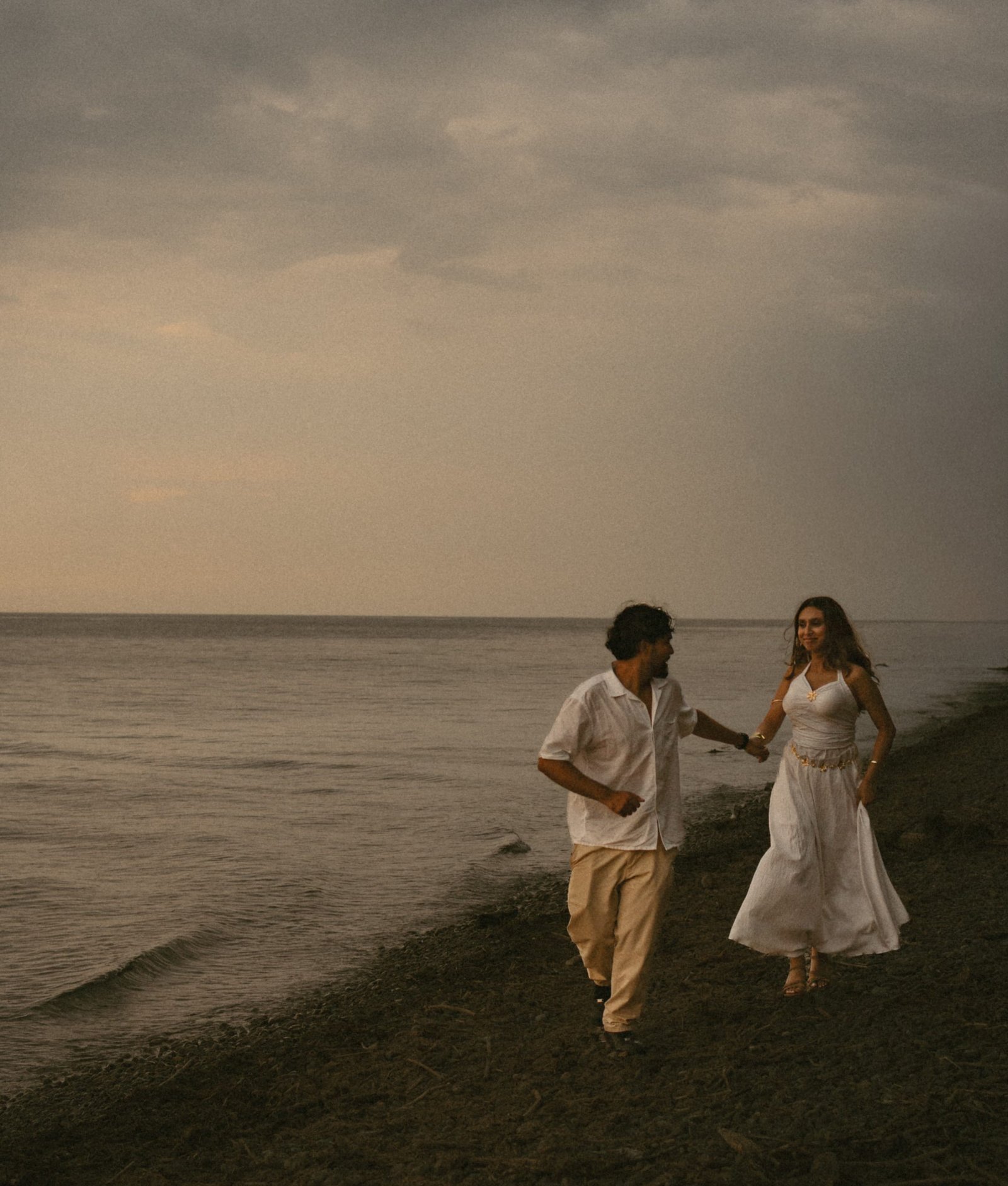 A couple walking hand in hand along an empty stony shoreline under a wide grey sky