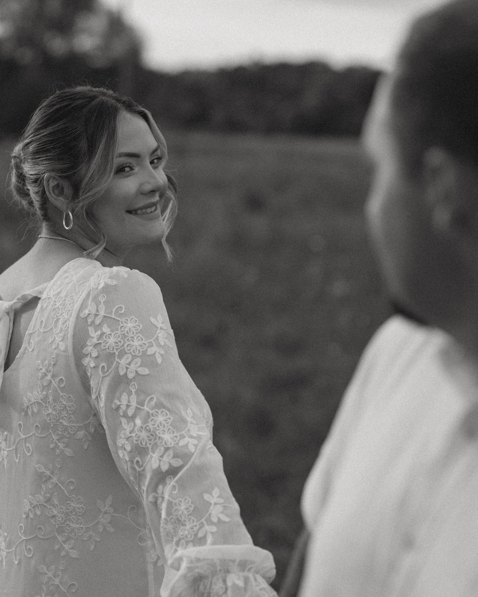 A black and white frame of a woman in a lace dress glancing back over her shoulder at her fiancé, smiling