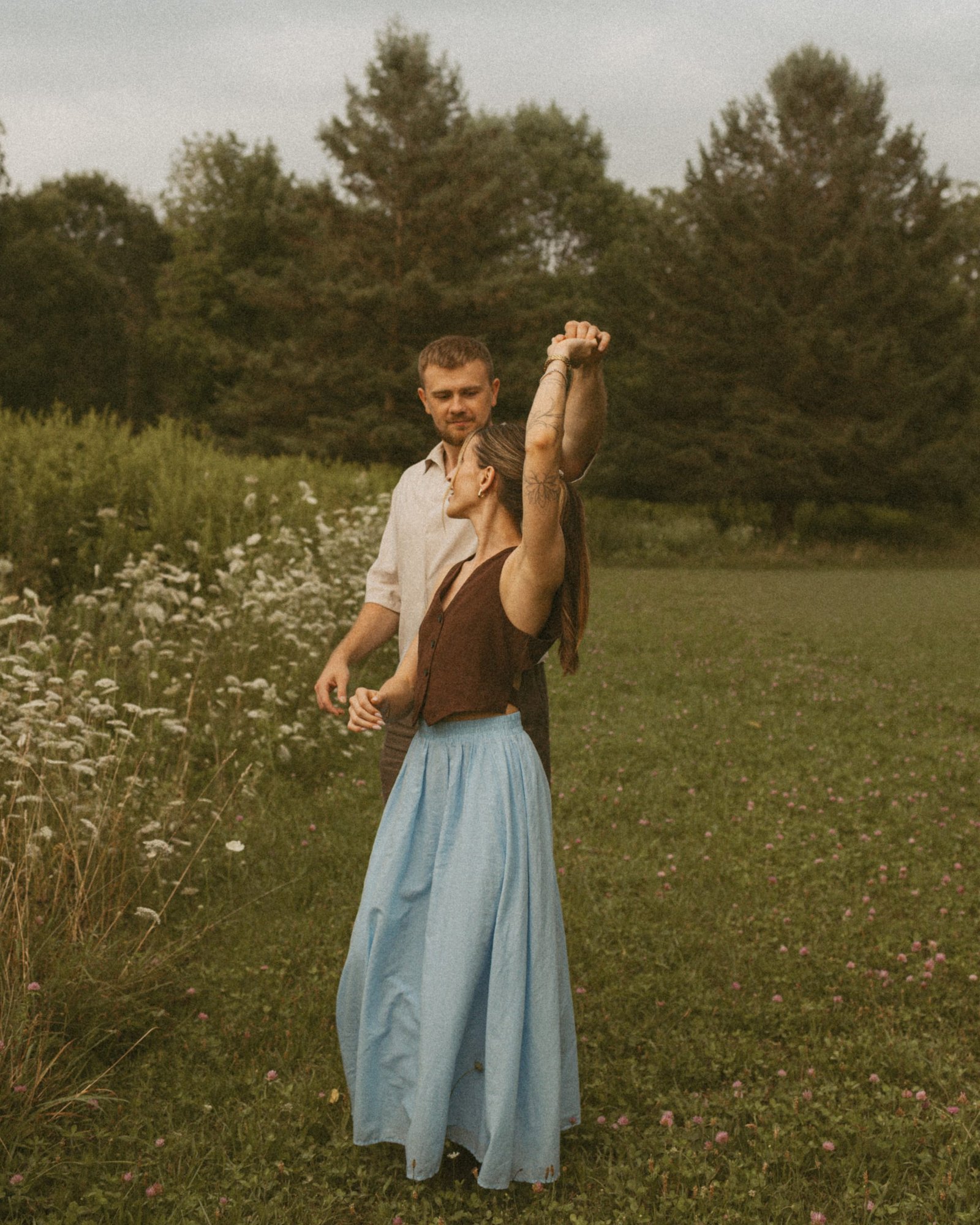 A man twirling his partner in a pale blue skirt across a green meadow by the lake