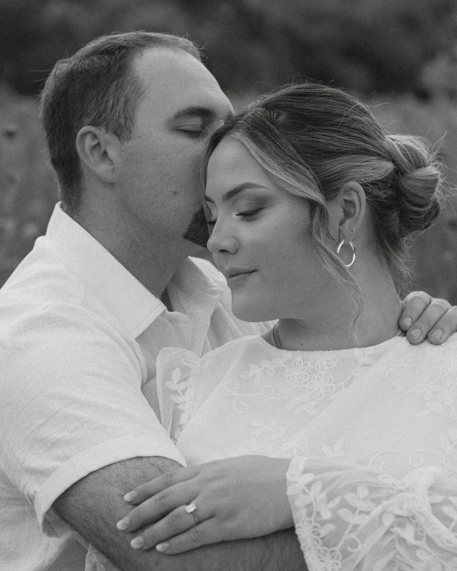 A black and white portrait of a man kissing his fiancée's temple as she holds his arm, eyes closed