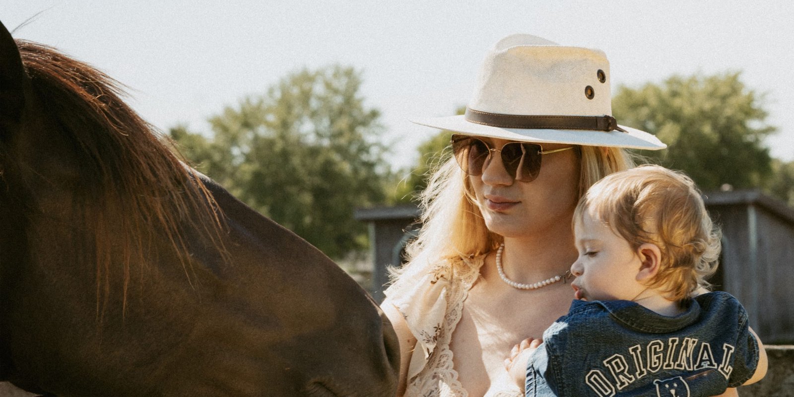 A mother in a wide-brim hat holding her toddler as a horse nuzzles close at a paddock fence