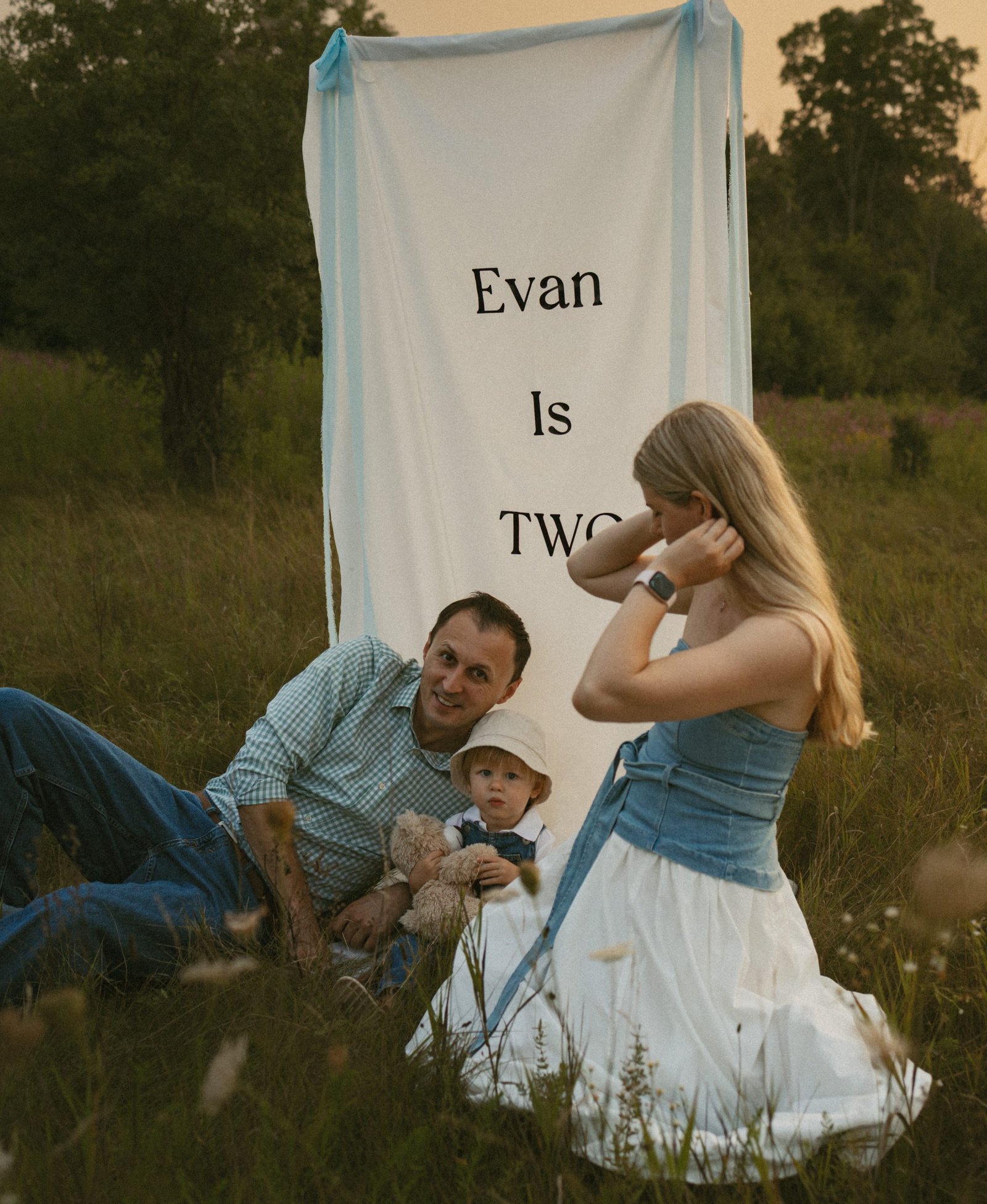 A mother, father and toddler sitting in a wildflower meadow beside a hanging fabric banner at sunset