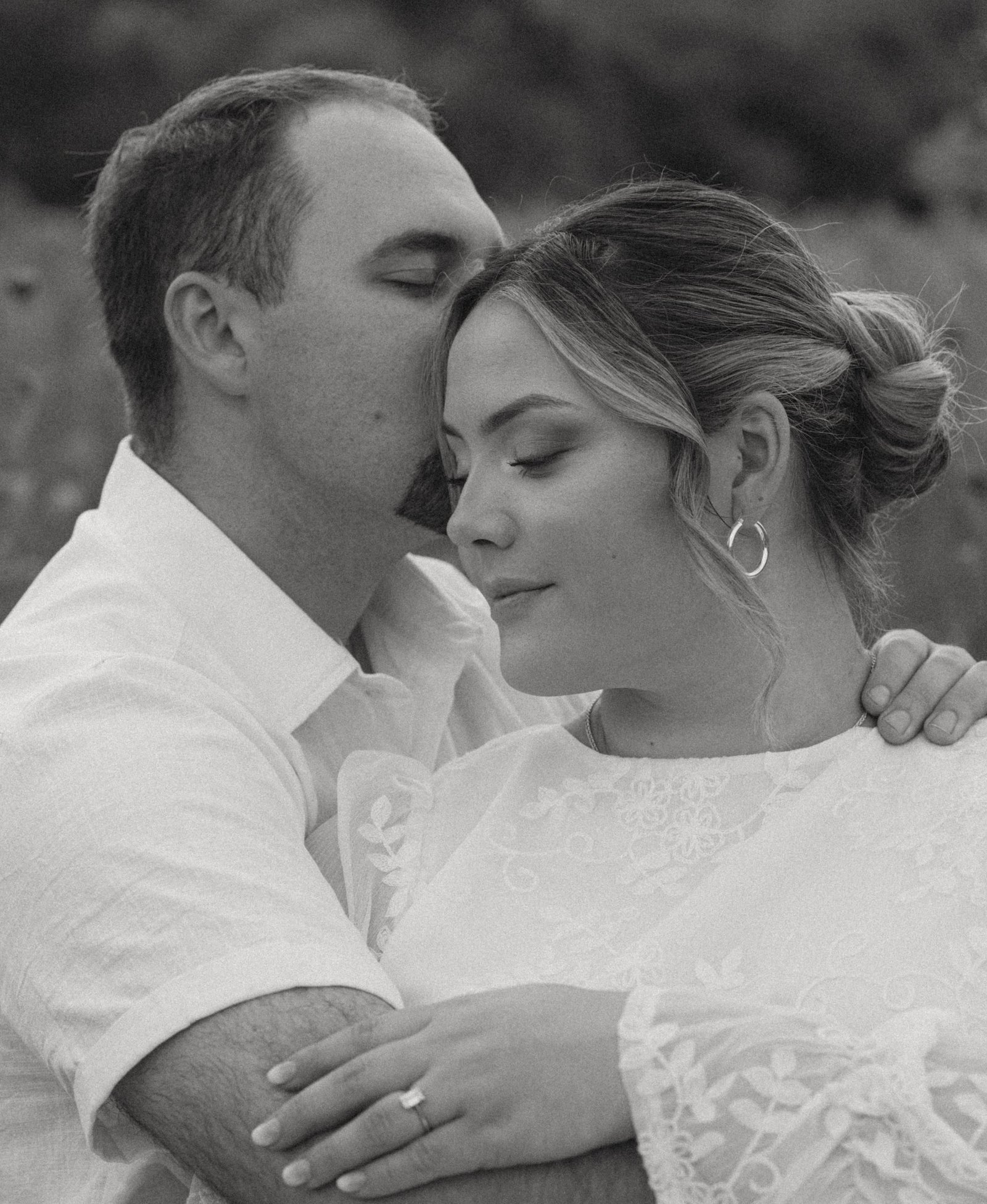A black and white portrait of a man kissing his fiancée's temple as she holds his arm, eyes closed