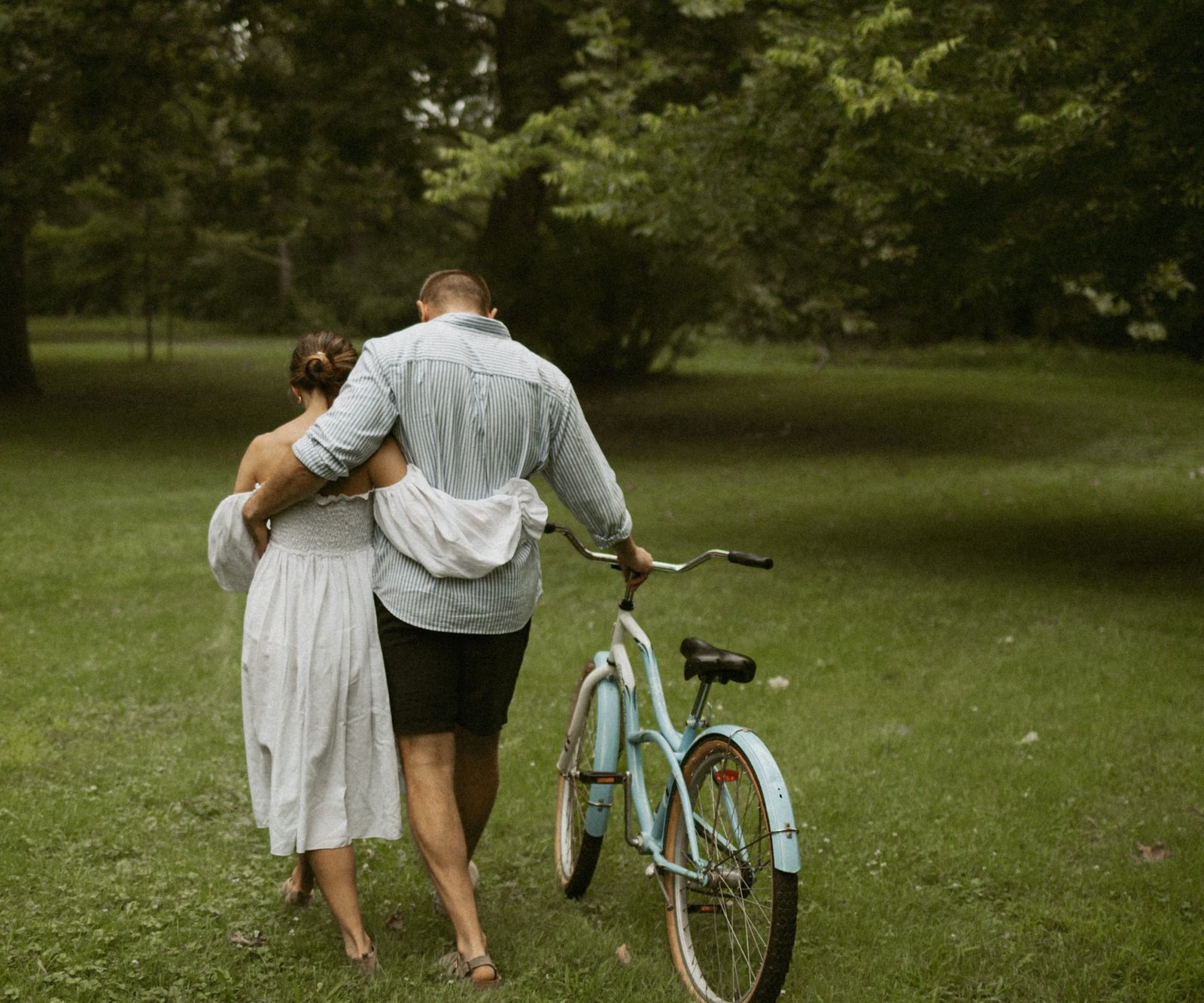 A couple walking hand in hand across a park lawn with a bicycle, seen from behind