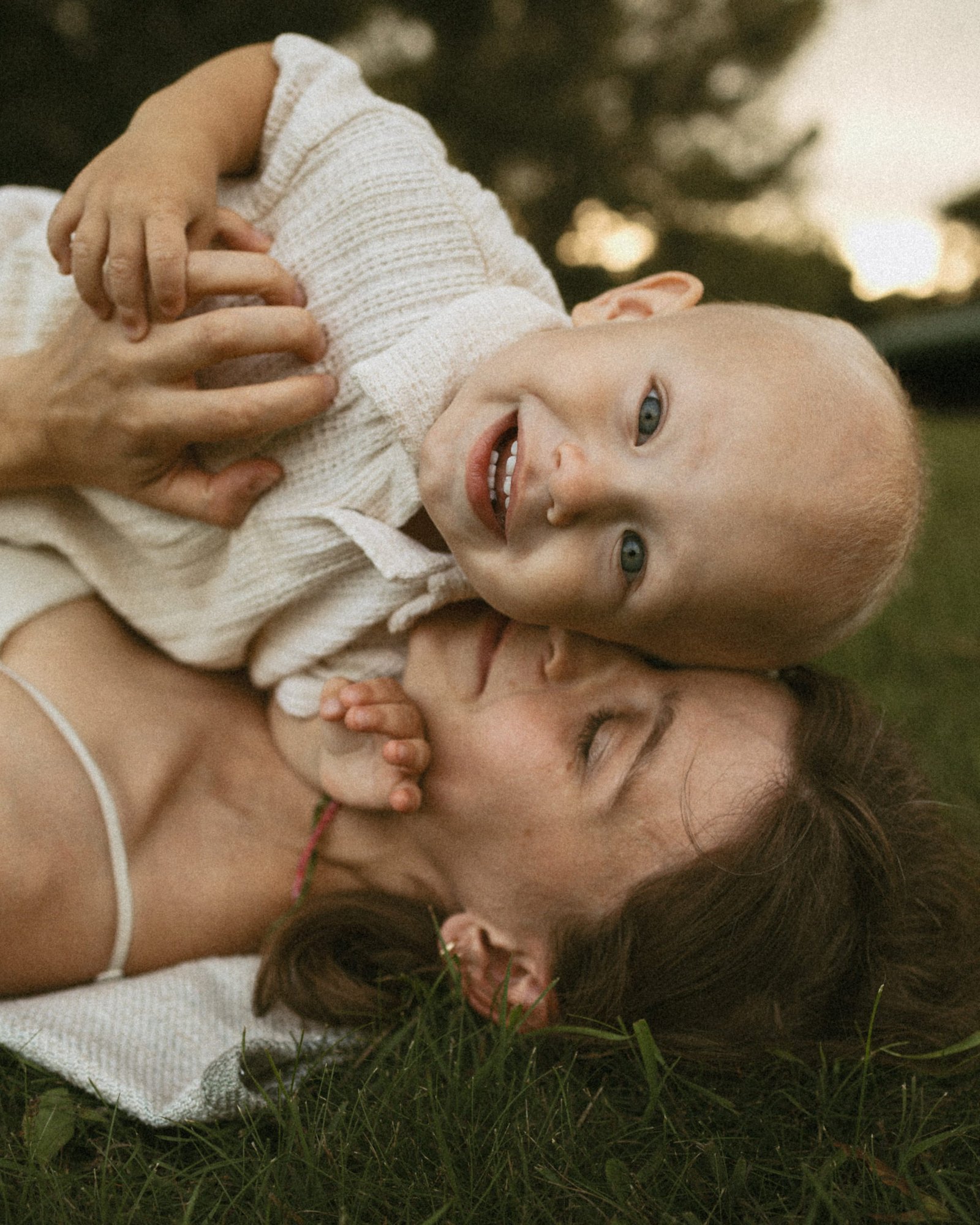 A laughing baby lying on top of his mother in the grass, pressing his cheek against hers