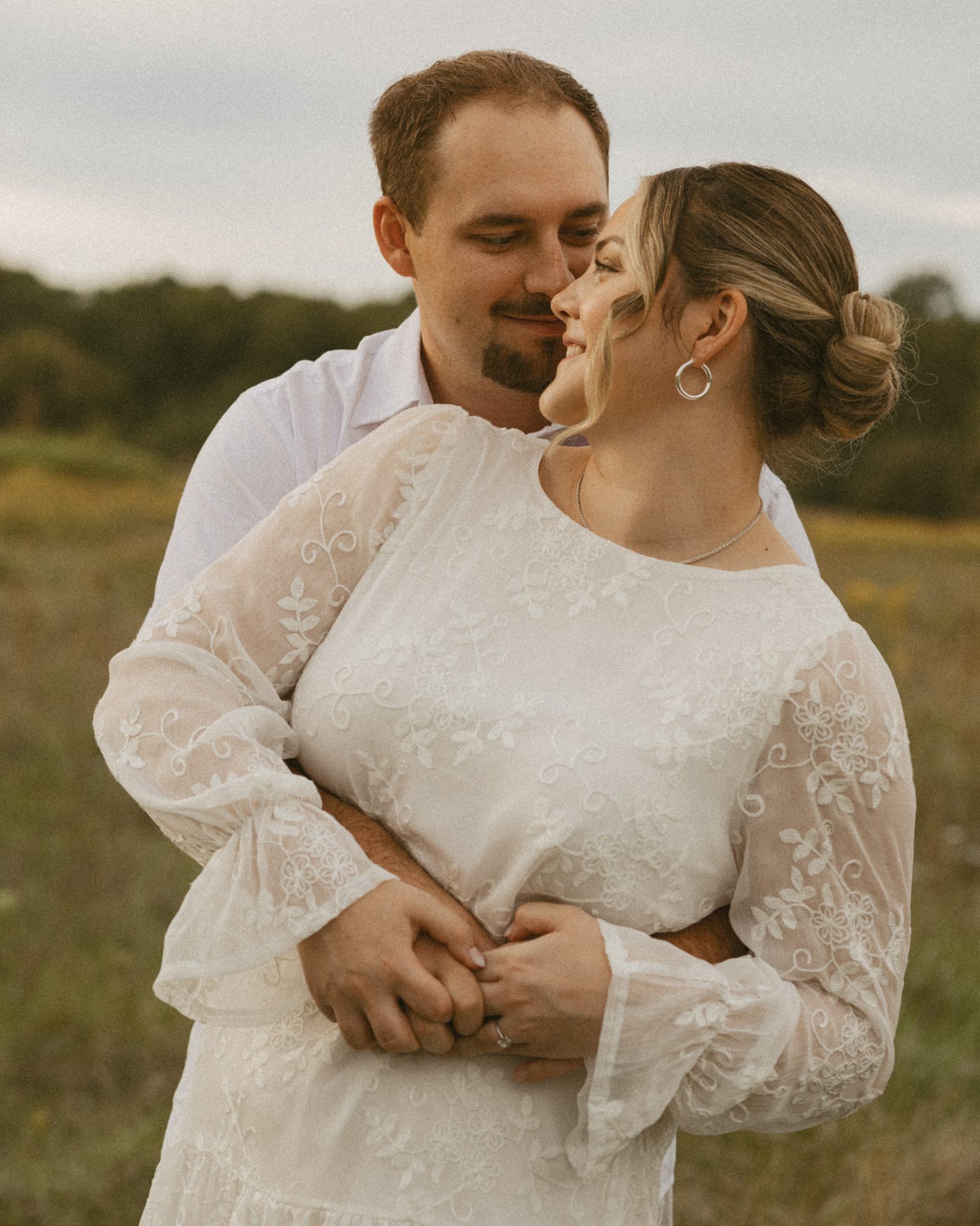 A man wrapping his arms around his fiancée from behind in a meadow, both of them smiling with eyes closed