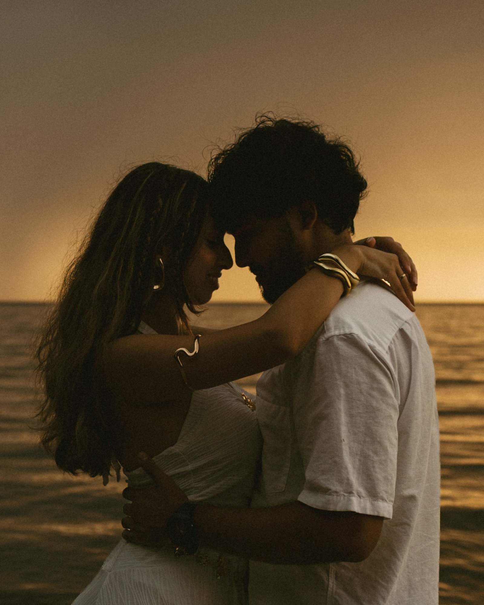 A couple with their foreheads touching at the water's edge in low orange light