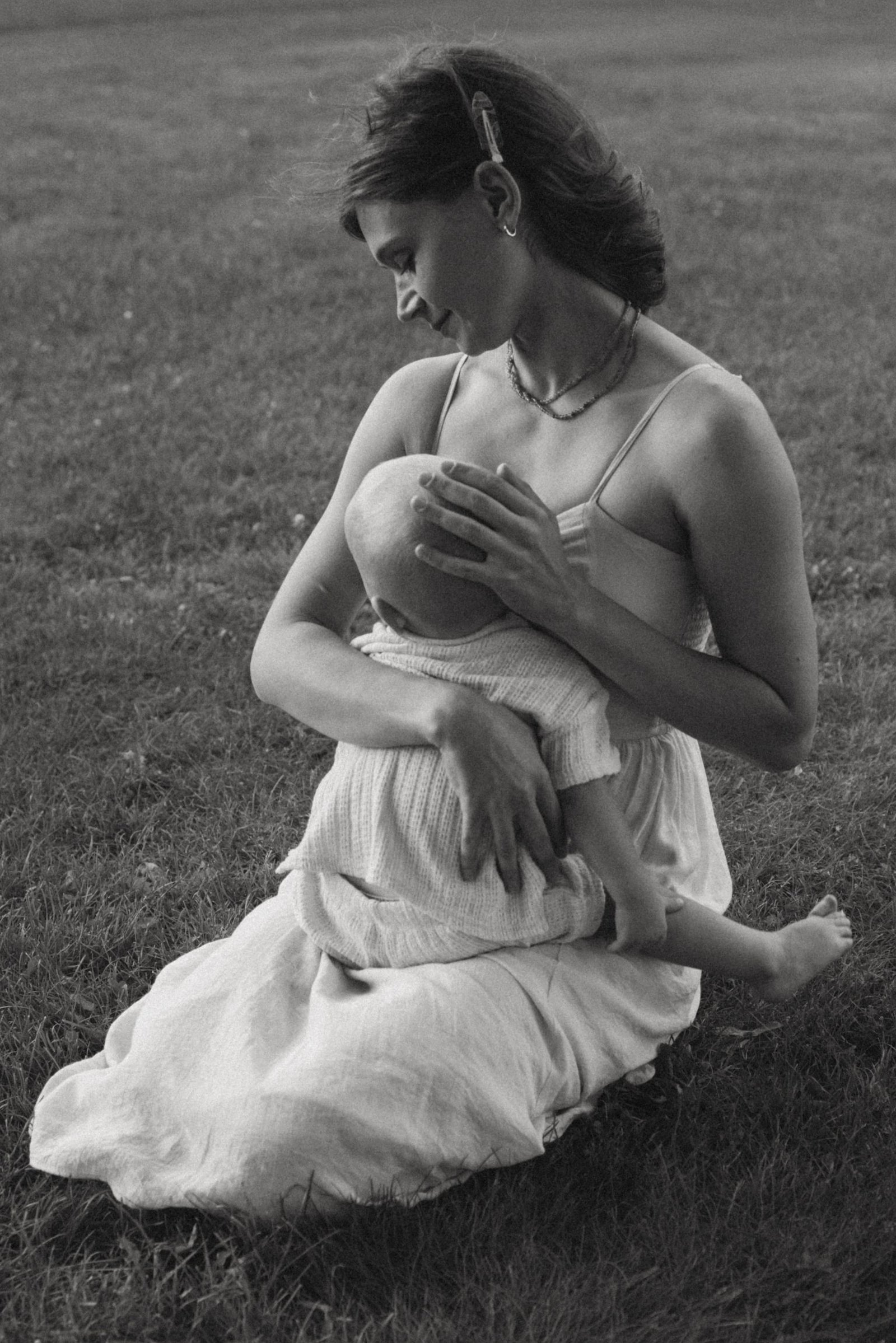 A black and white frame of a mother kneeling in the grass, breastfeeding her son