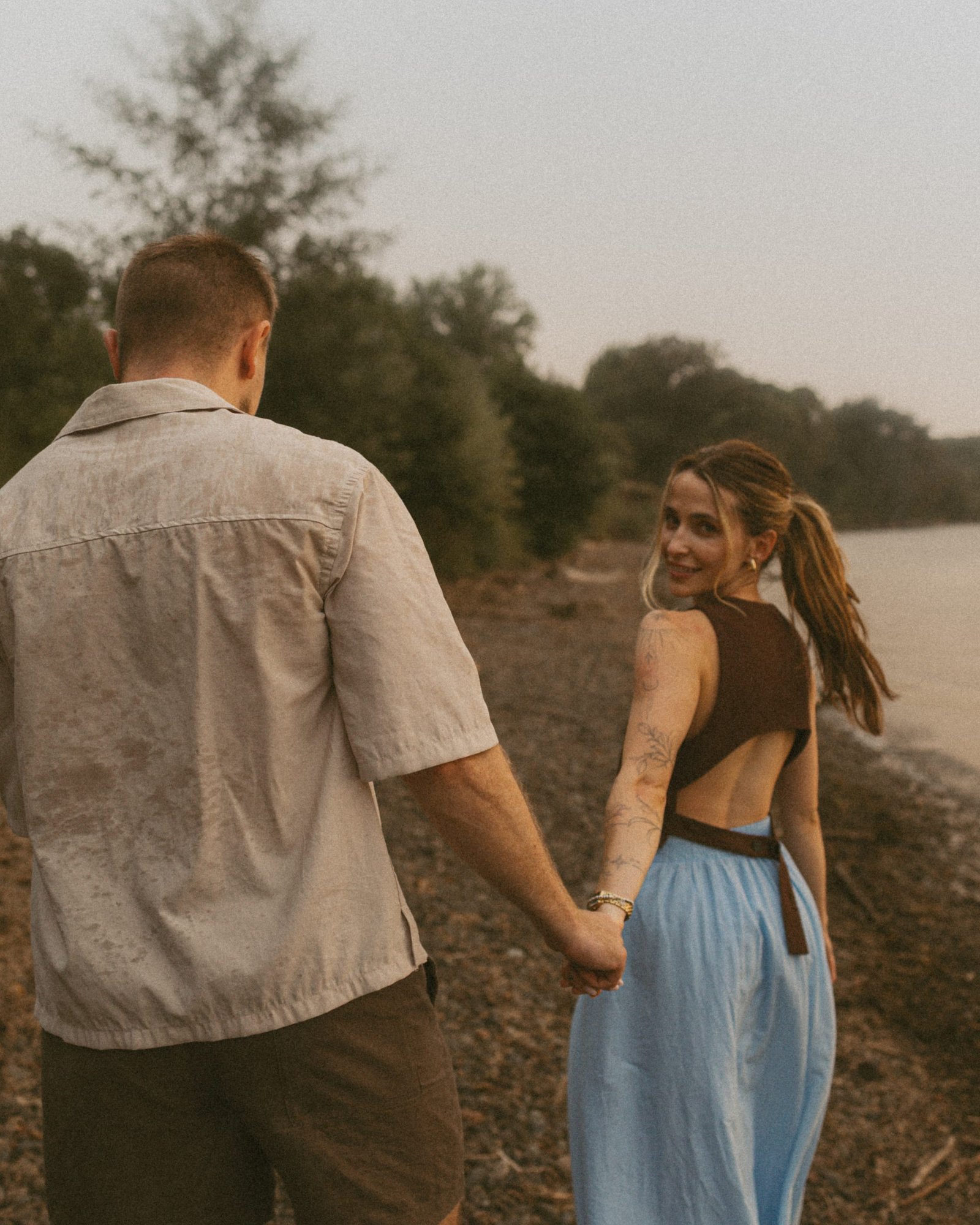A couple walking hand in hand along a pebble beach on Lake Ontario, she looks back over her shoulder and smiles