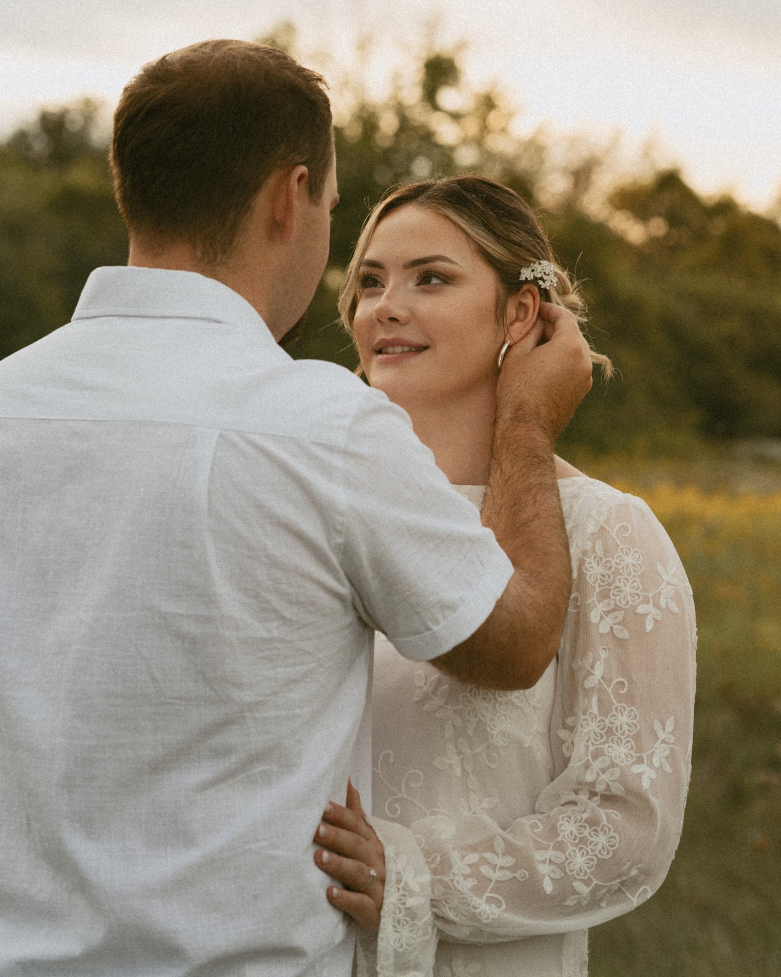 A woman with a wildflower in her hair smiling at the camera while her fiancé holds her close in the meadow