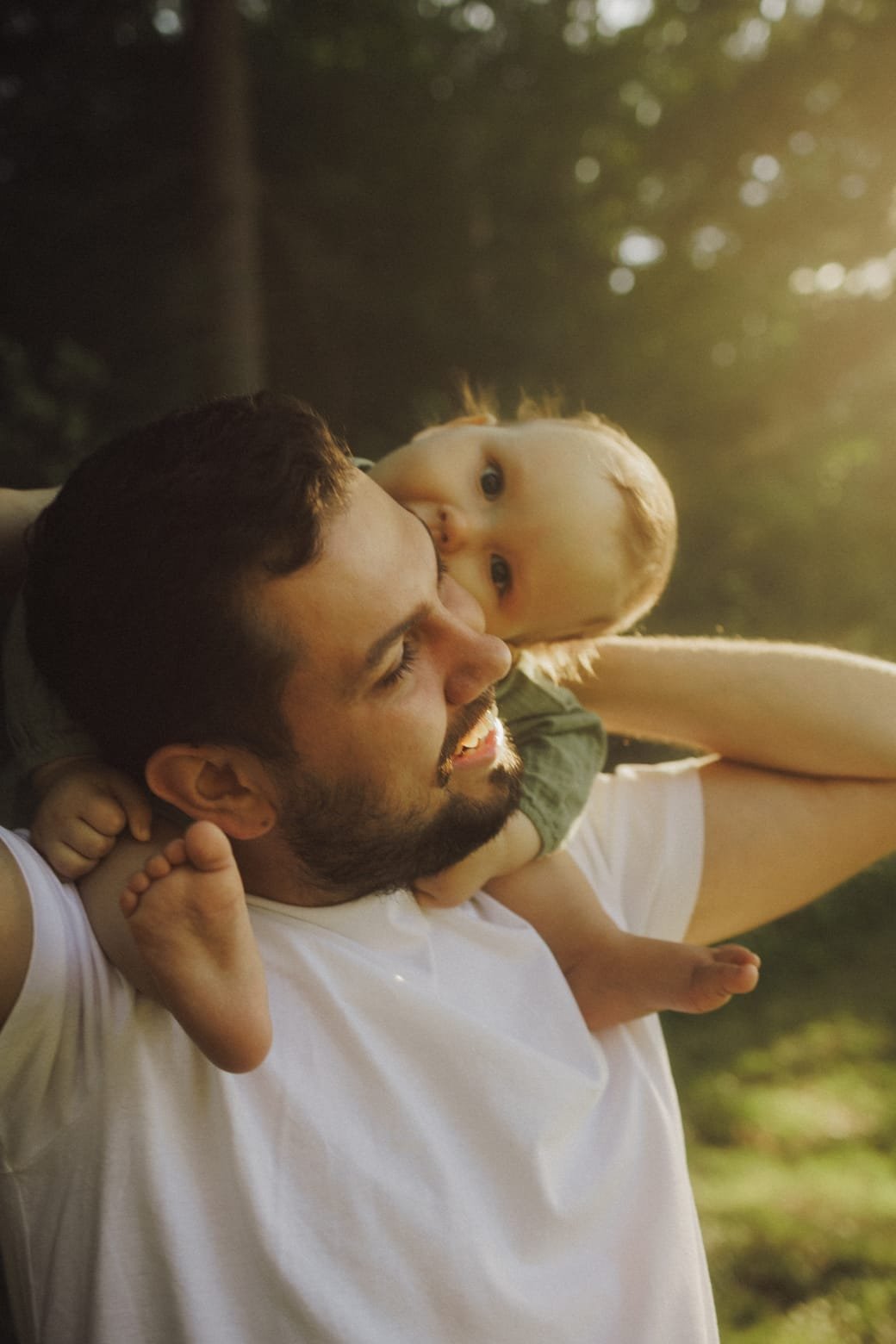 A baby looking back over his father's shoulder on a shaded forest path