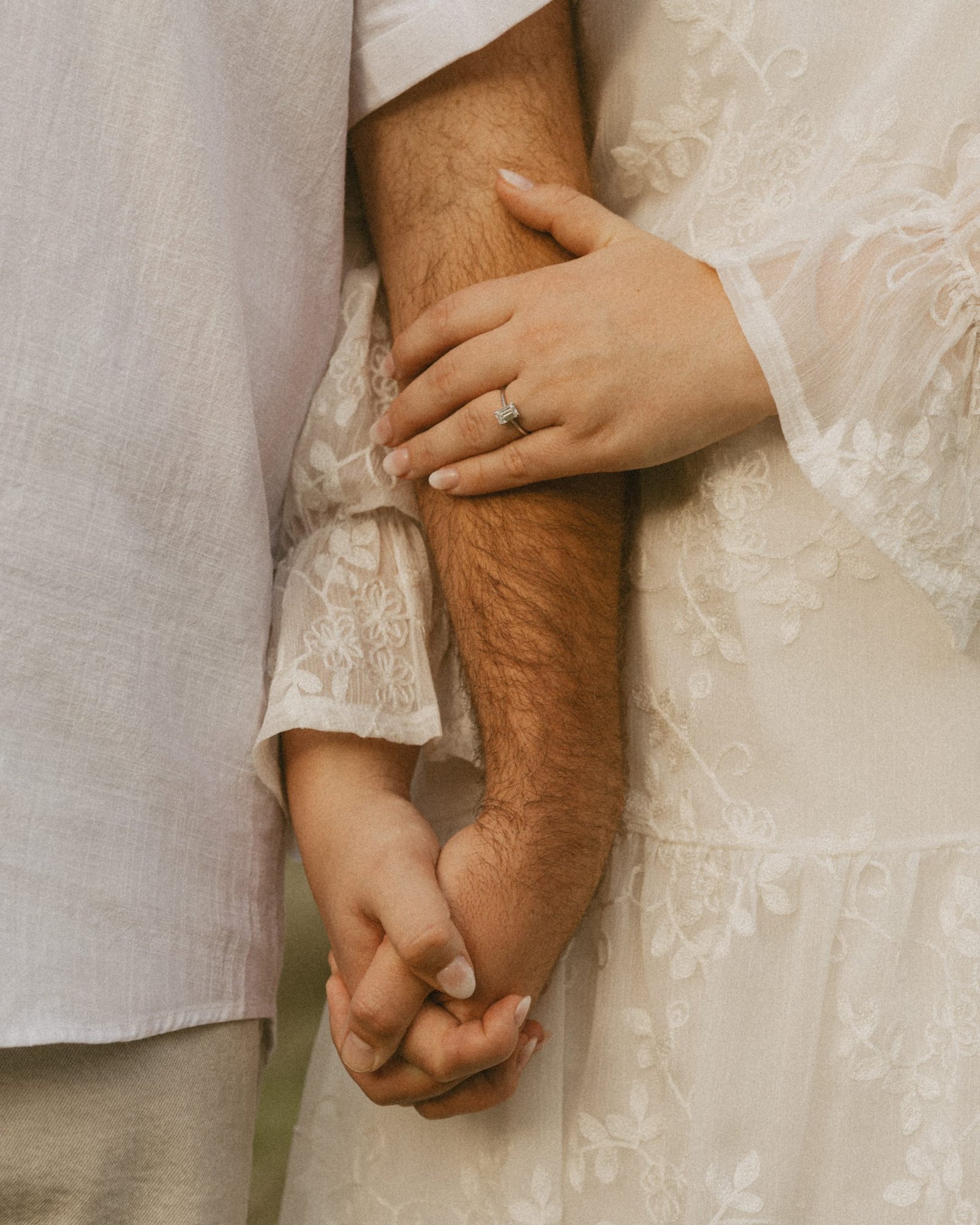 Close detail of a woman's hand with an engagement ring resting on her fiancé's arm, fingers laced together