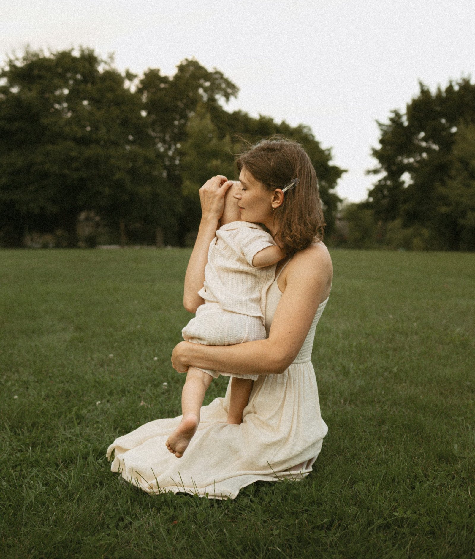 A mother kneeling on a wide park lawn, holding her son close, tall trees behind them