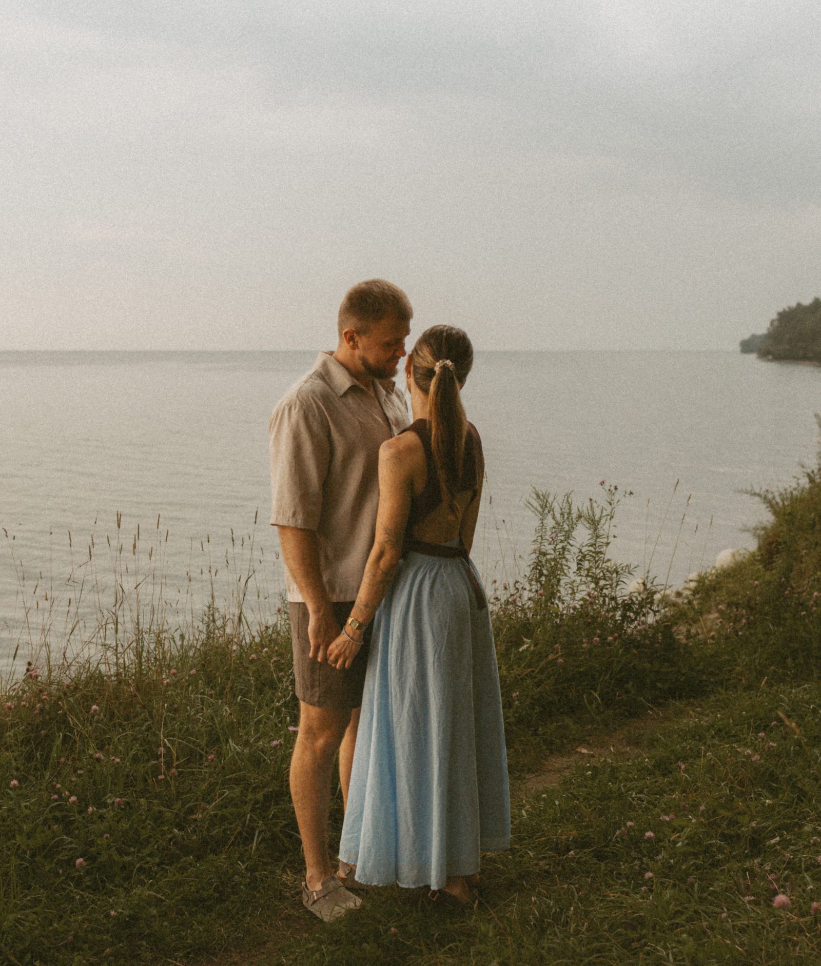 A couple standing close on a grassy bluff above Lake Ontario under a heavy grey sky