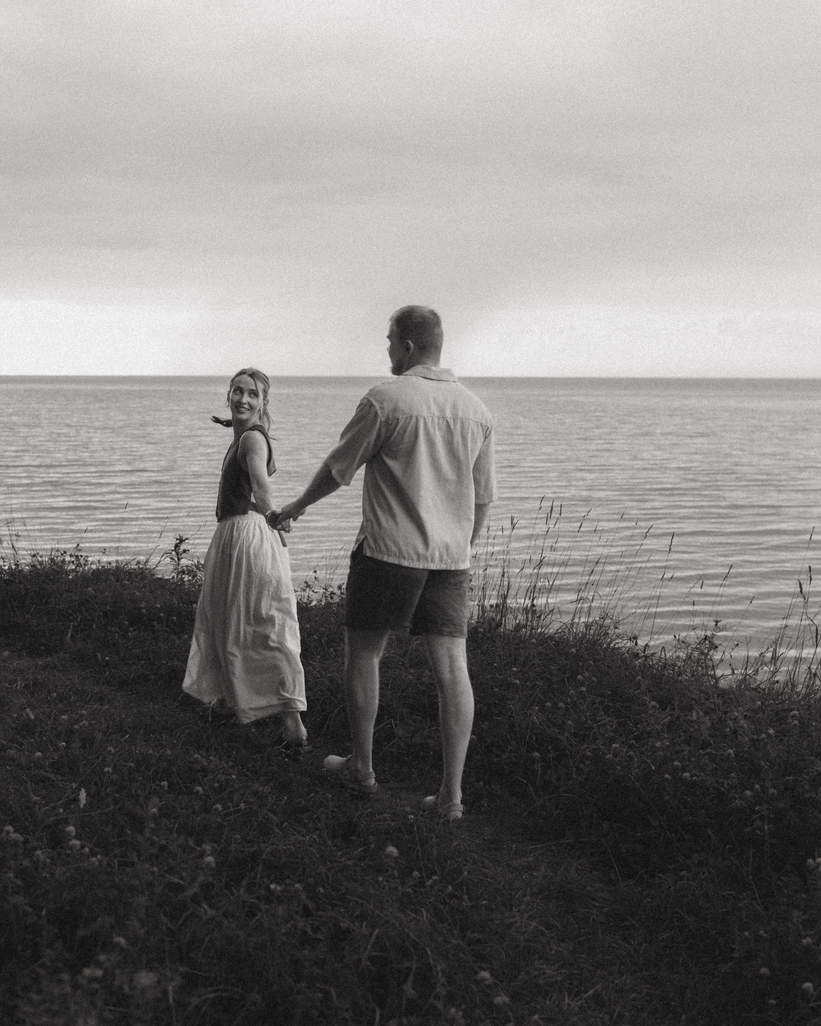 A black and white frame of a couple walking hand in hand toward the edge of the bluff and the open lake