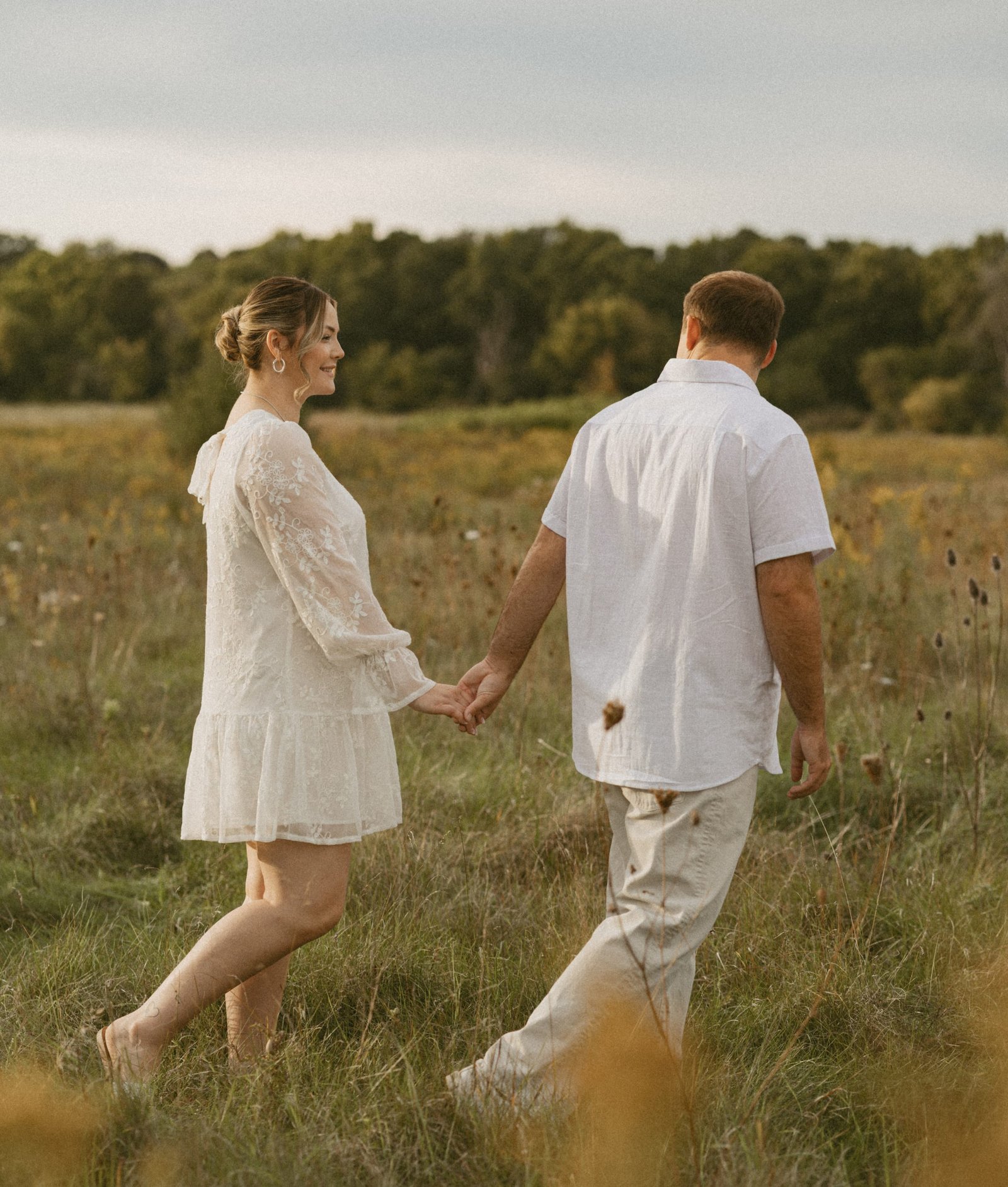 A couple in white walking hand in hand into a wildflower meadow, a line of trees behind them