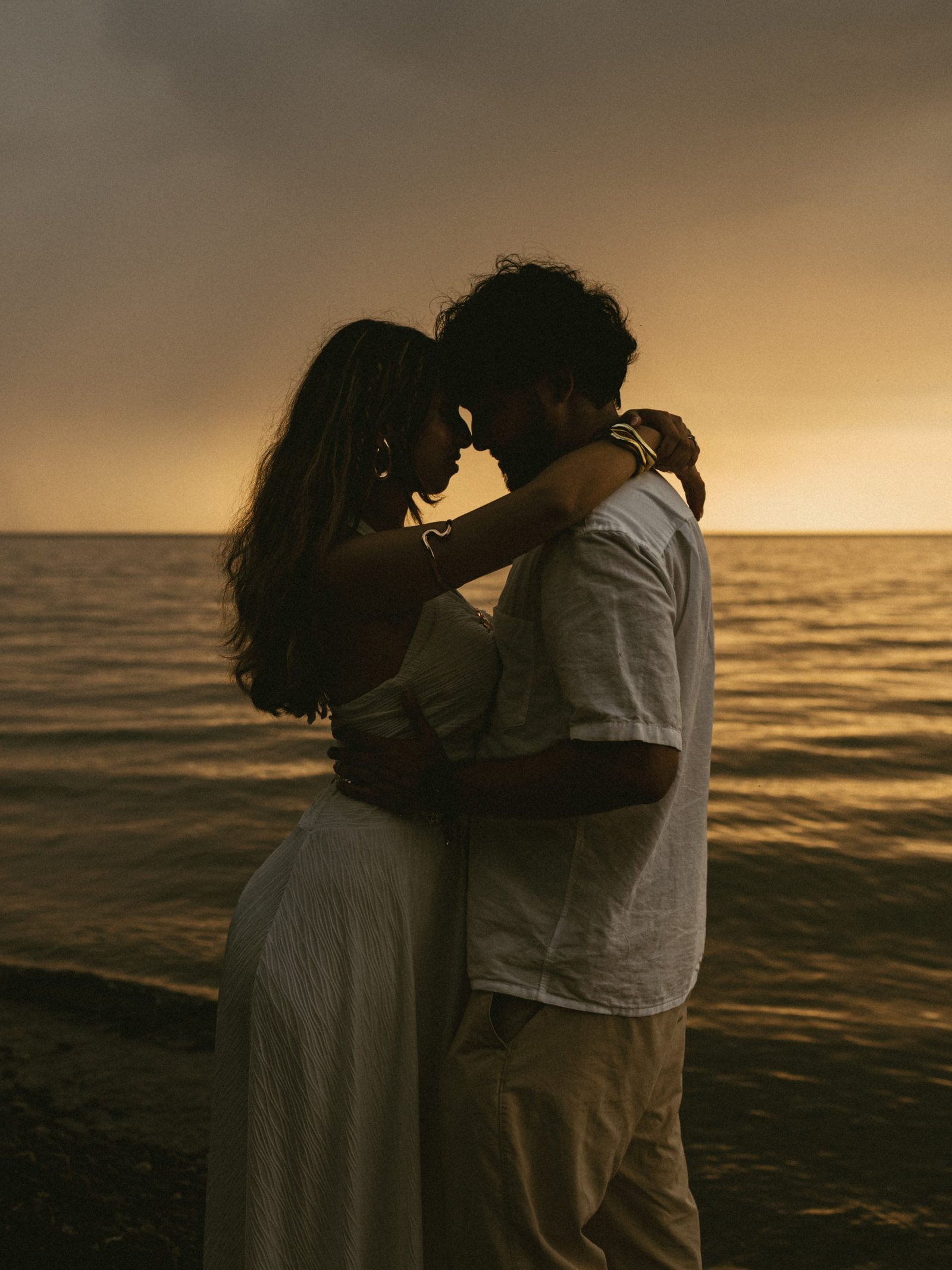 A couple standing close together on a stony lakeshore as the sun sets behind heavy cloud
