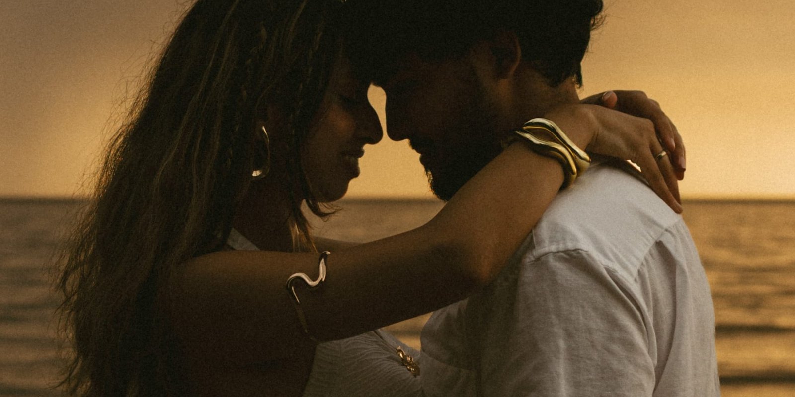 A couple with their foreheads touching at the water's edge in low orange light