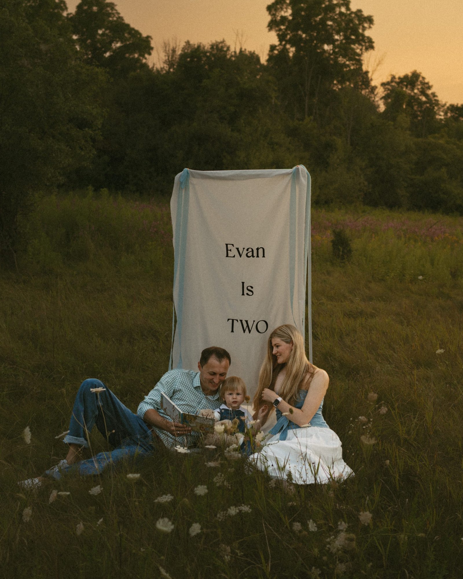 A family of three seated on the grass in front of a linen banner reading Evan Is Two