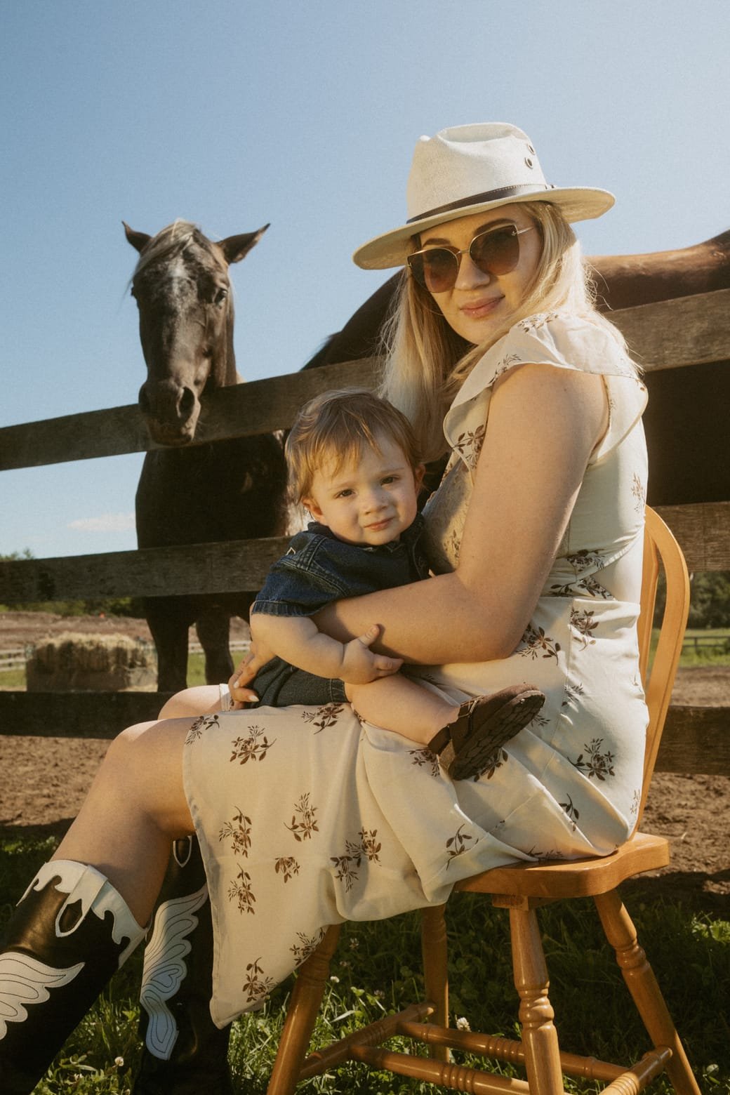 A mother in a wide-brim hat holding her toddler son on her lap beside a horse paddock