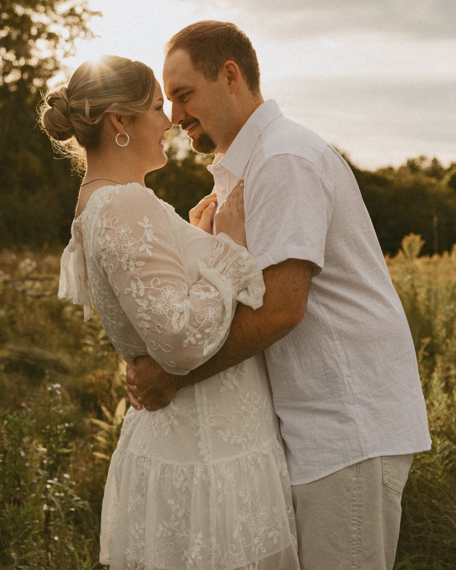 A couple in white holding each other close in a tall-grass meadow, the low sun glowing through the trees behind them