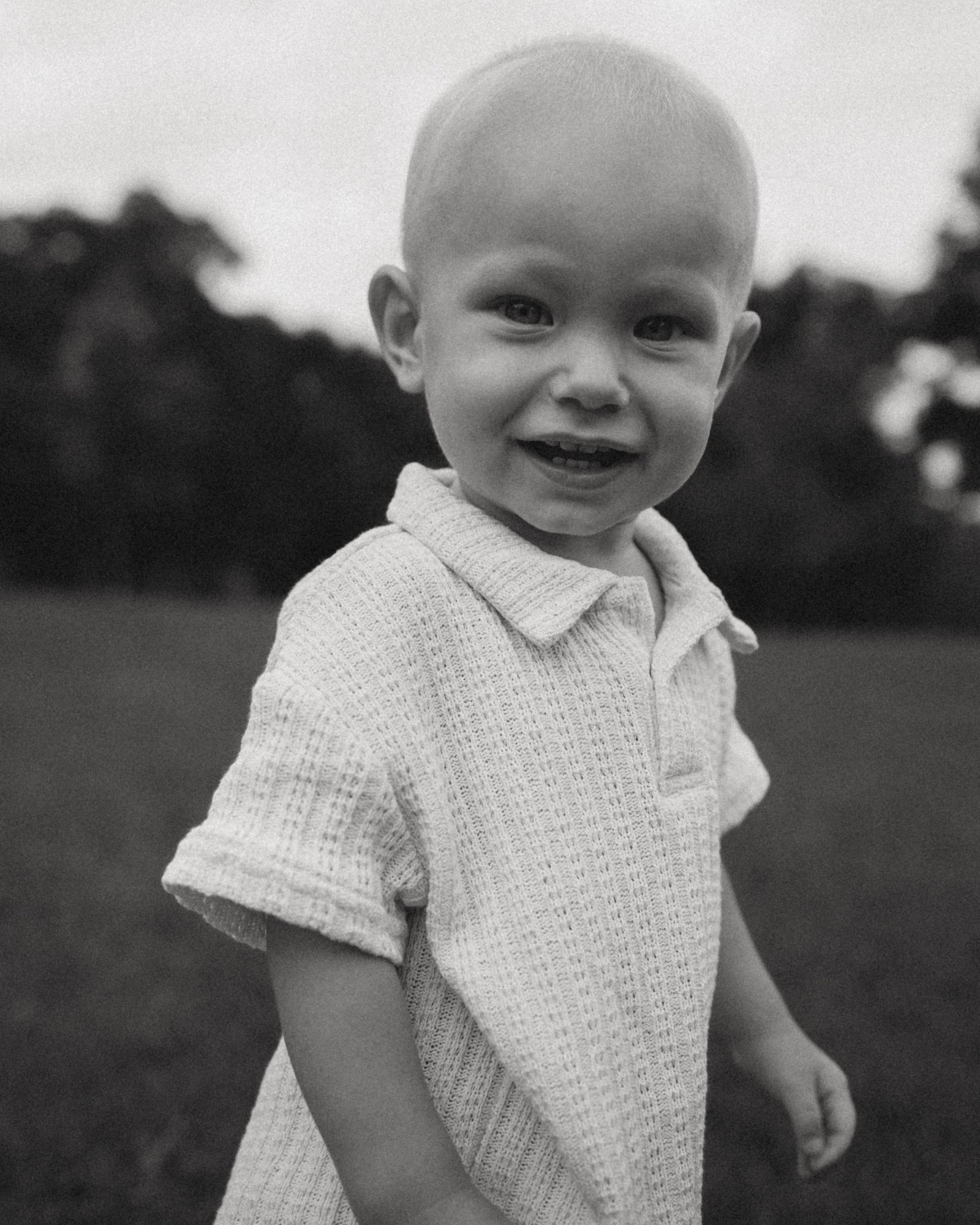 A black and white portrait of a grinning baby boy in a linen shirt standing in the park
