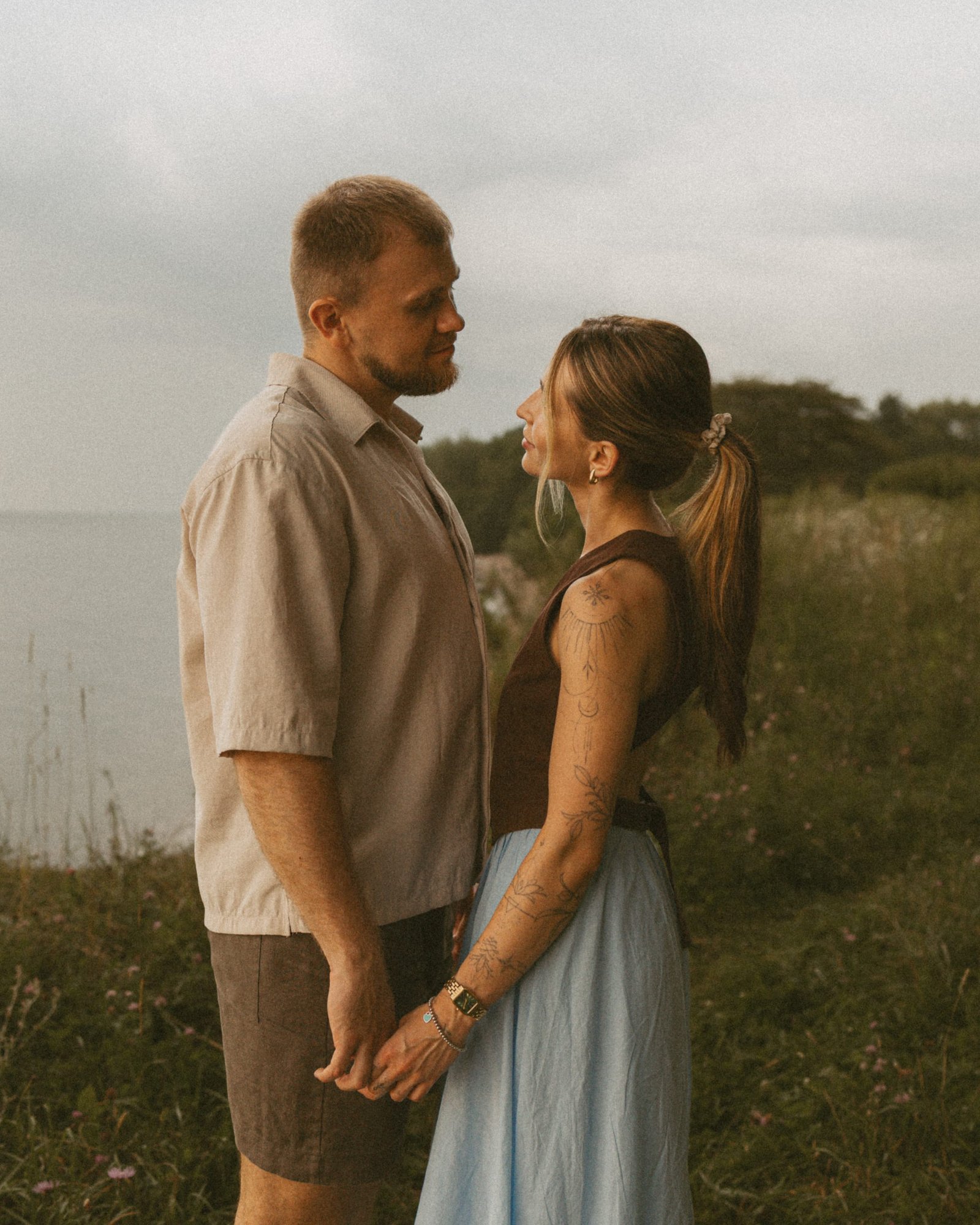 A couple standing face to face holding hands on the lakeshore, wildflowers and the grey water behind them
