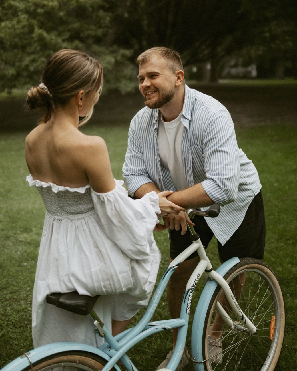 A woman in a white off-shoulder dress steadying a vintage blue bicycle while her partner holds her