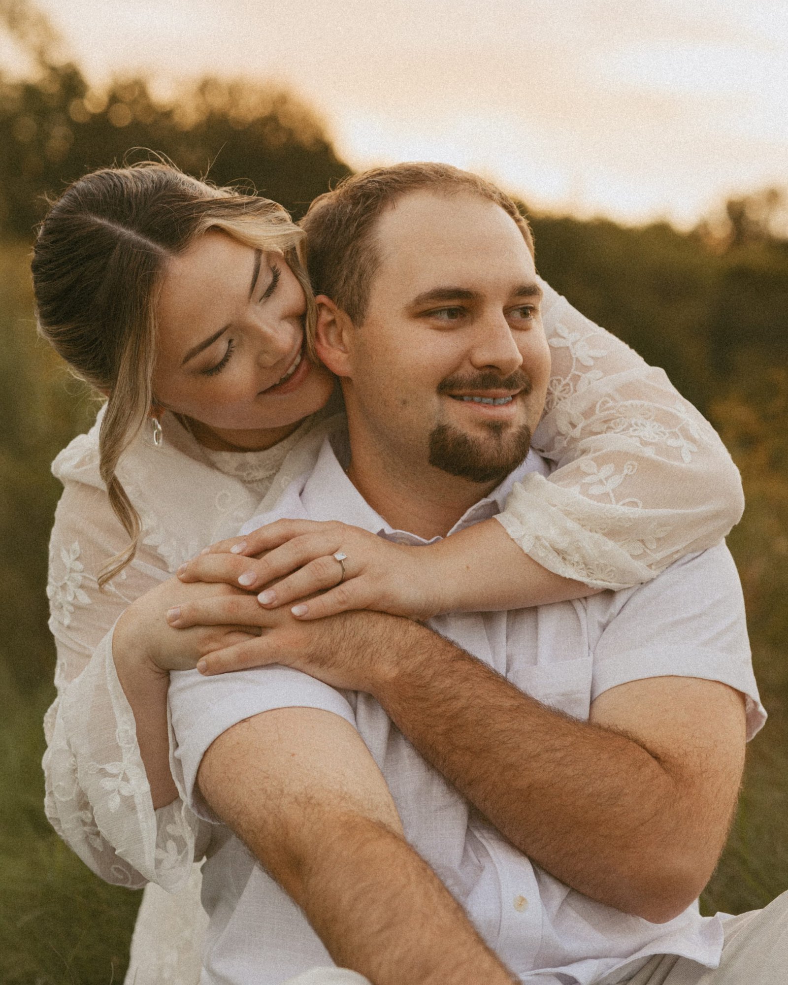 A woman hugging her fiancé from behind, cheek pressed to his, both of them grinning in the meadow