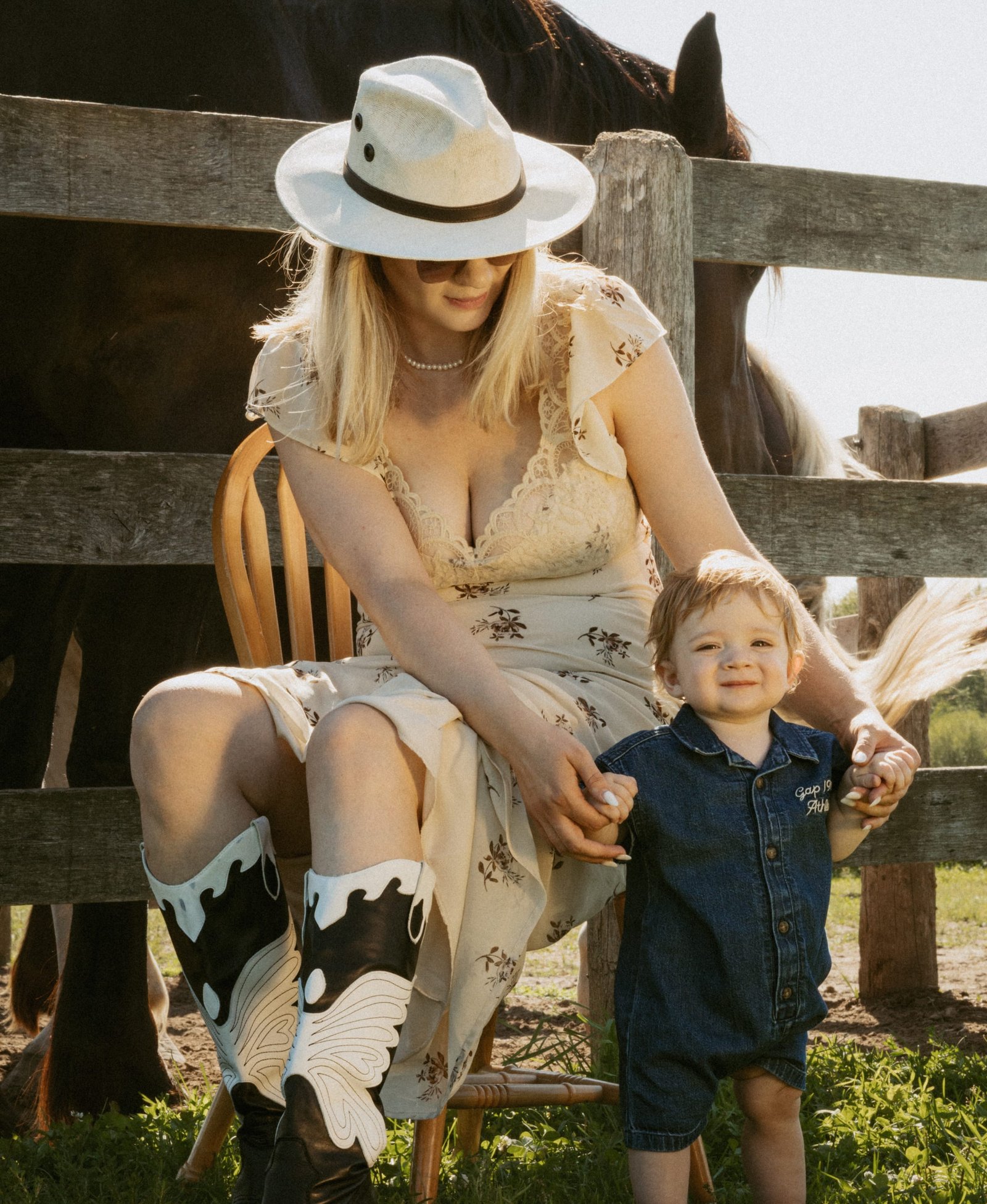 A woman in a floral dress and cowboy boots standing at a paddock fence with her son on her hip