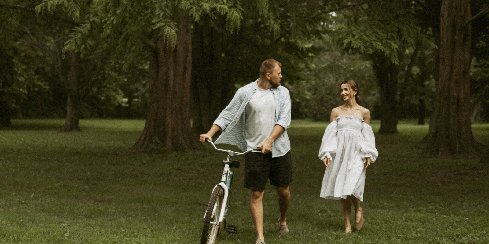 A couple walking together across a park lawn, the man wheeling a vintage bicycle beside her