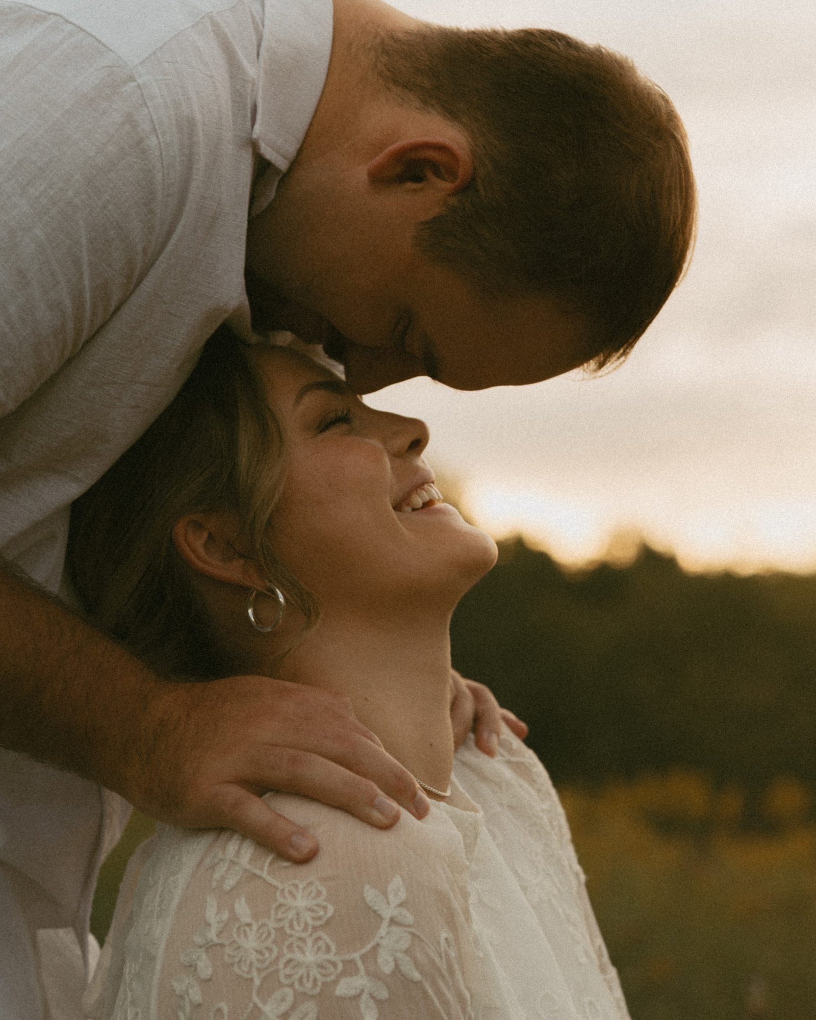 A man leaning down to kiss his fiancée's forehead as she laughs, sunset behind them